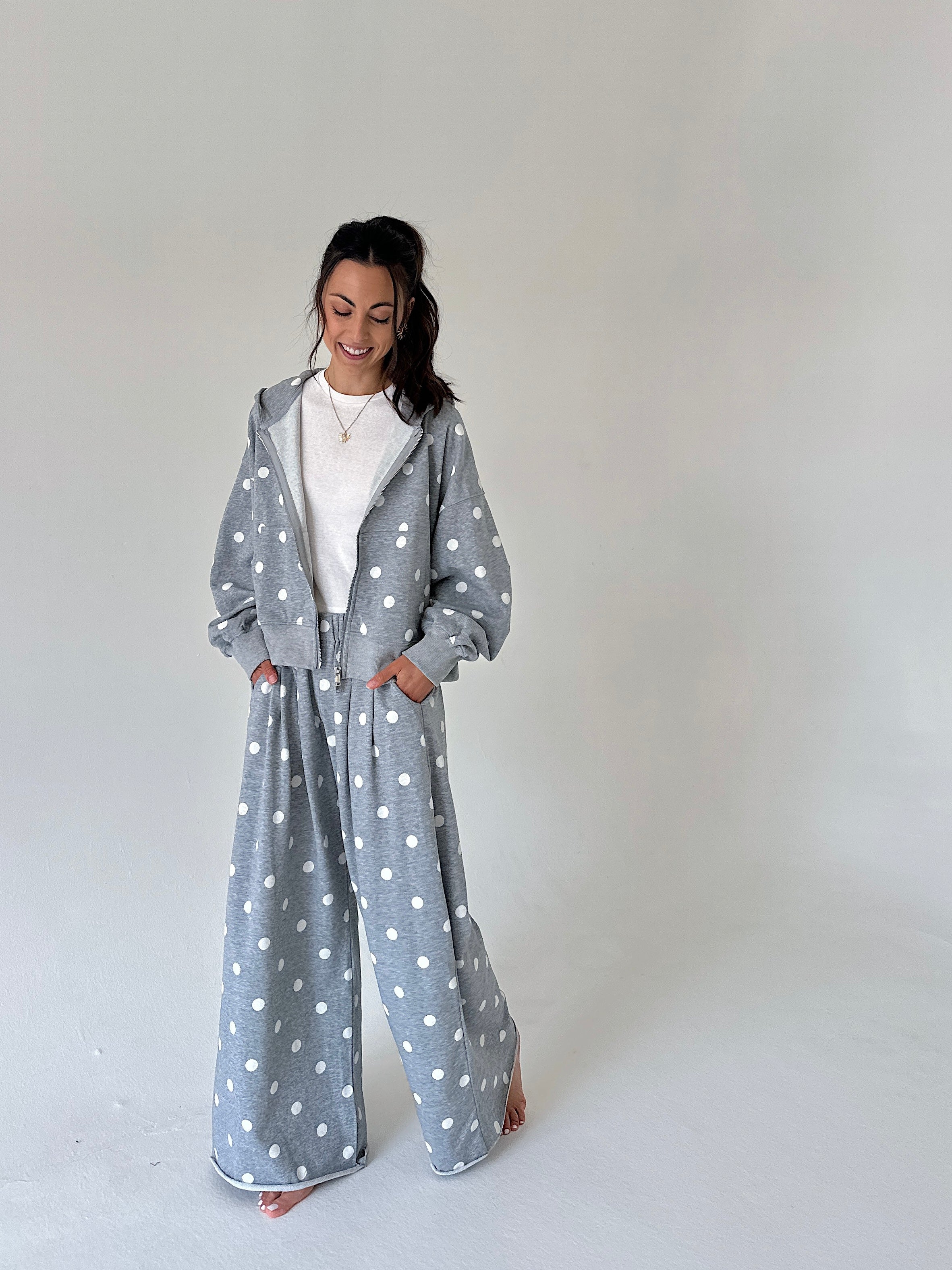 A woman modeling a light gray two-piece loungewear set with white polka dots, consisting of a zip-up hoodie and matching wide-leg pants, styled over a white t-shirt against a plain light gray studio background.
