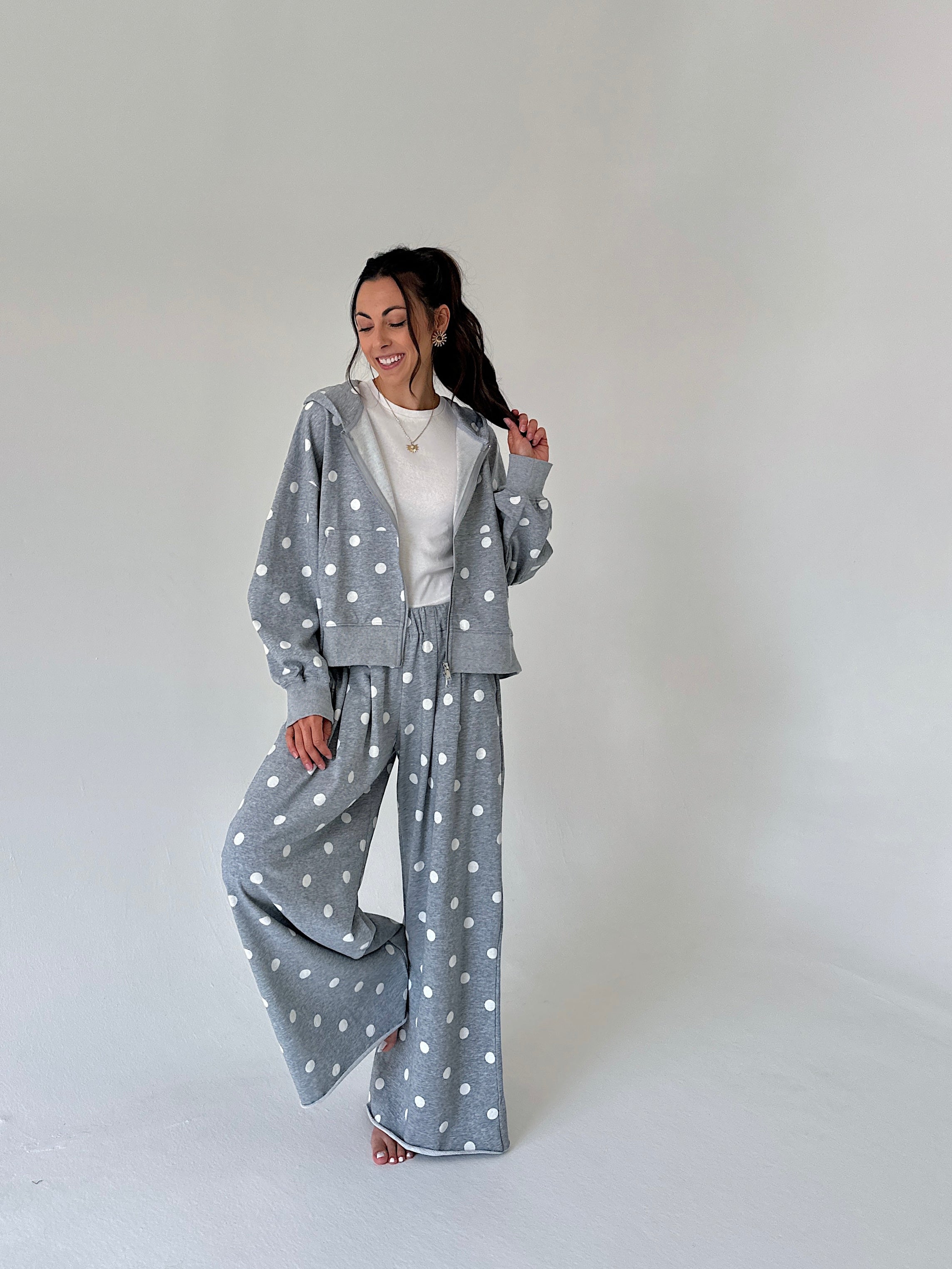 A woman modeling a gray two-piece loungewear set with white polka dots, consisting of a cropped zip-up hoodie and matching wide-leg pants, styled over a white t-shirt in a studio setting.
