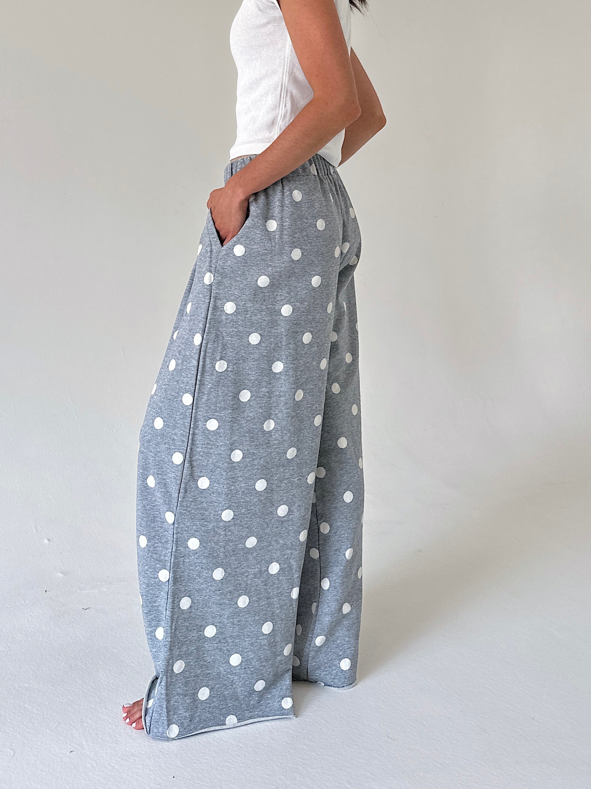 A person wearing wide-leg gray pants with a white polka dot pattern, paired with a white sleeveless top, standing against a plain light gray studio background.
