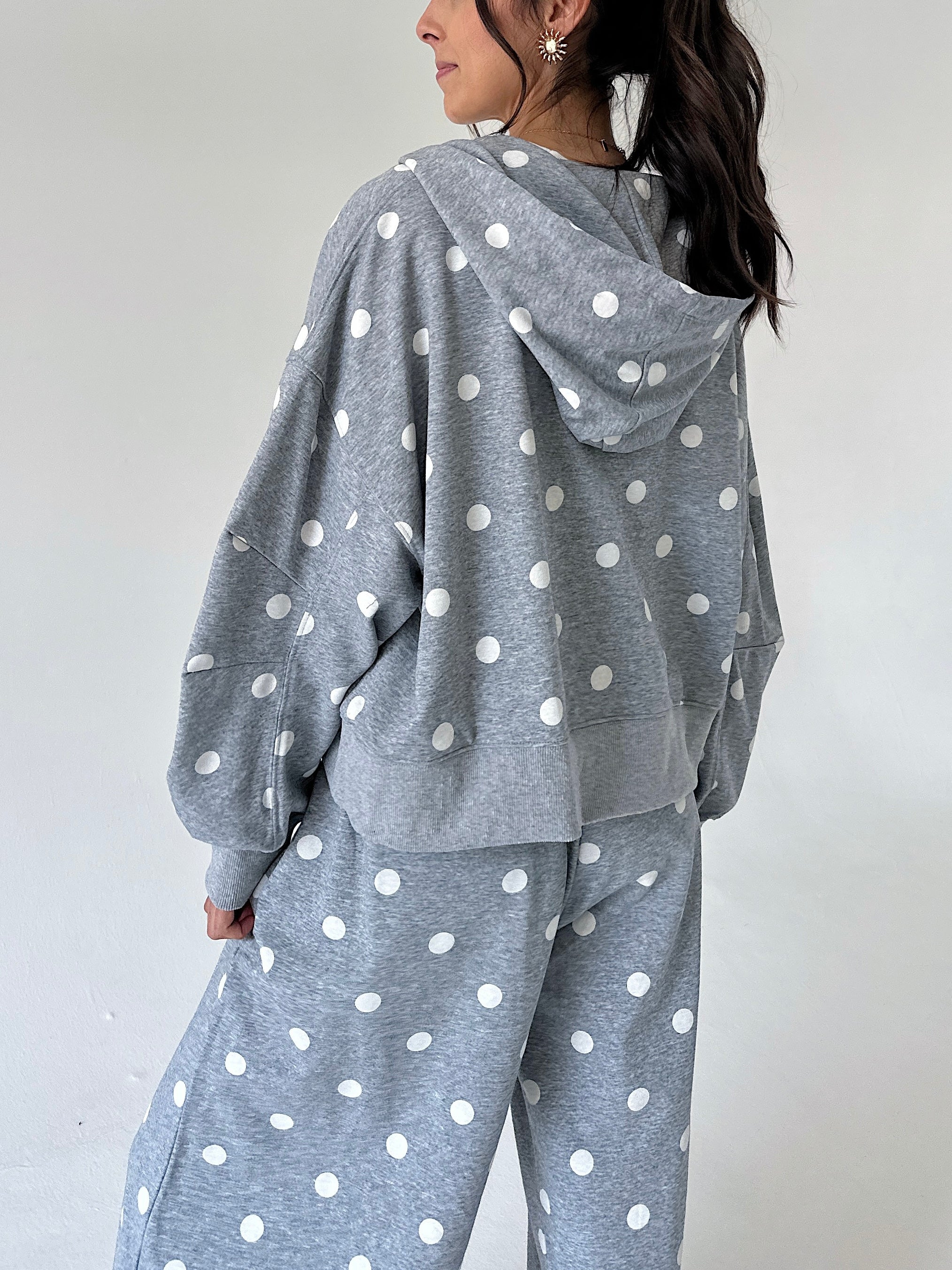 A woman wearing a heather gray hooded sweatshirt and matching sweatpants with a white polka dot pattern, shown from a side-back angle against a plain light gray wall.