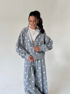 A woman wearing a matching gray and white polka dot lounge set, consisting of a cropped zip-up hoodie and matching sweatpants, standing against a plain off-white wall.