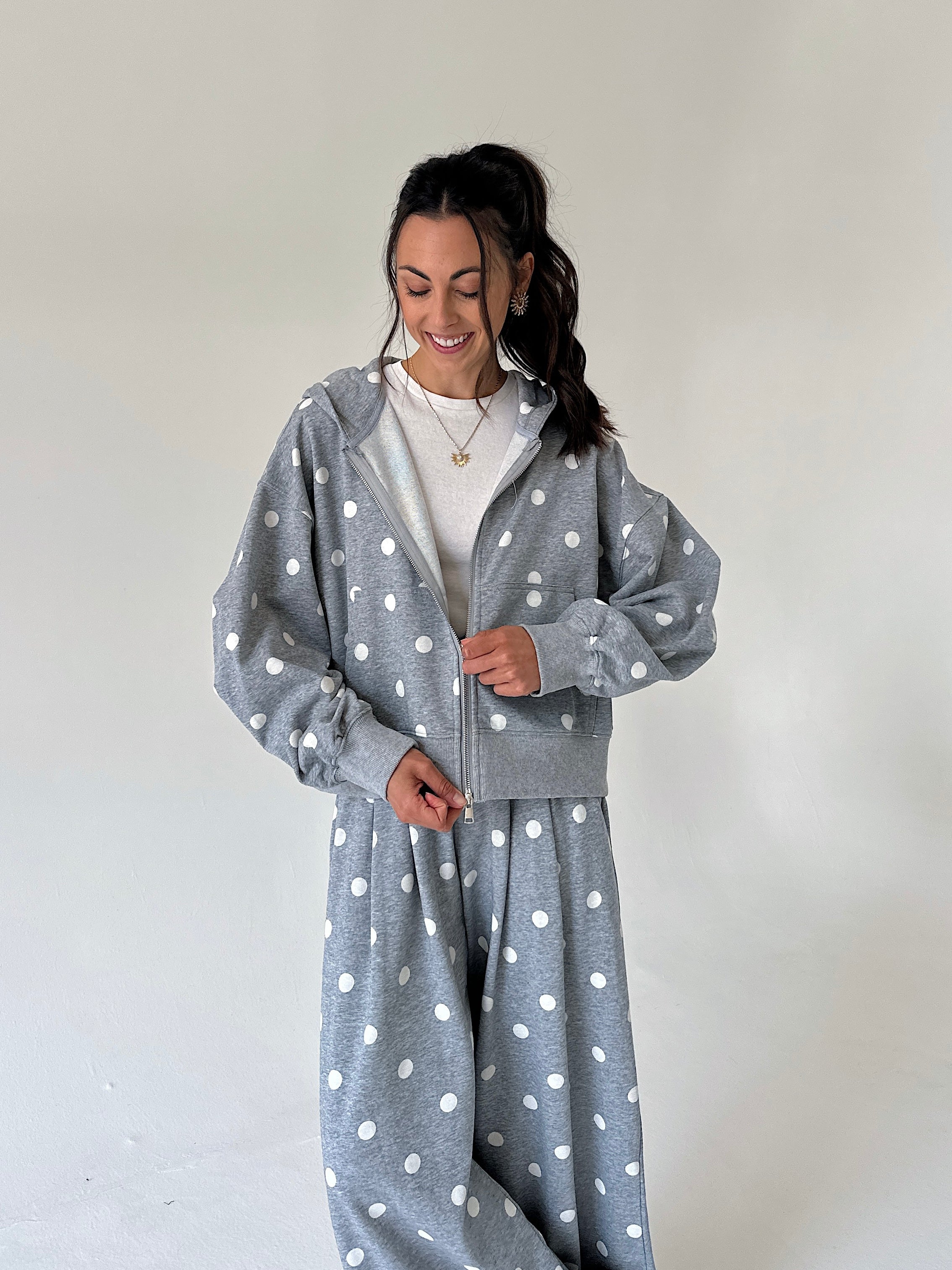 A woman wearing a matching gray and white polka dot lounge set, consisting of a cropped zip-up hoodie and matching sweatpants, standing against a plain off-white wall.
