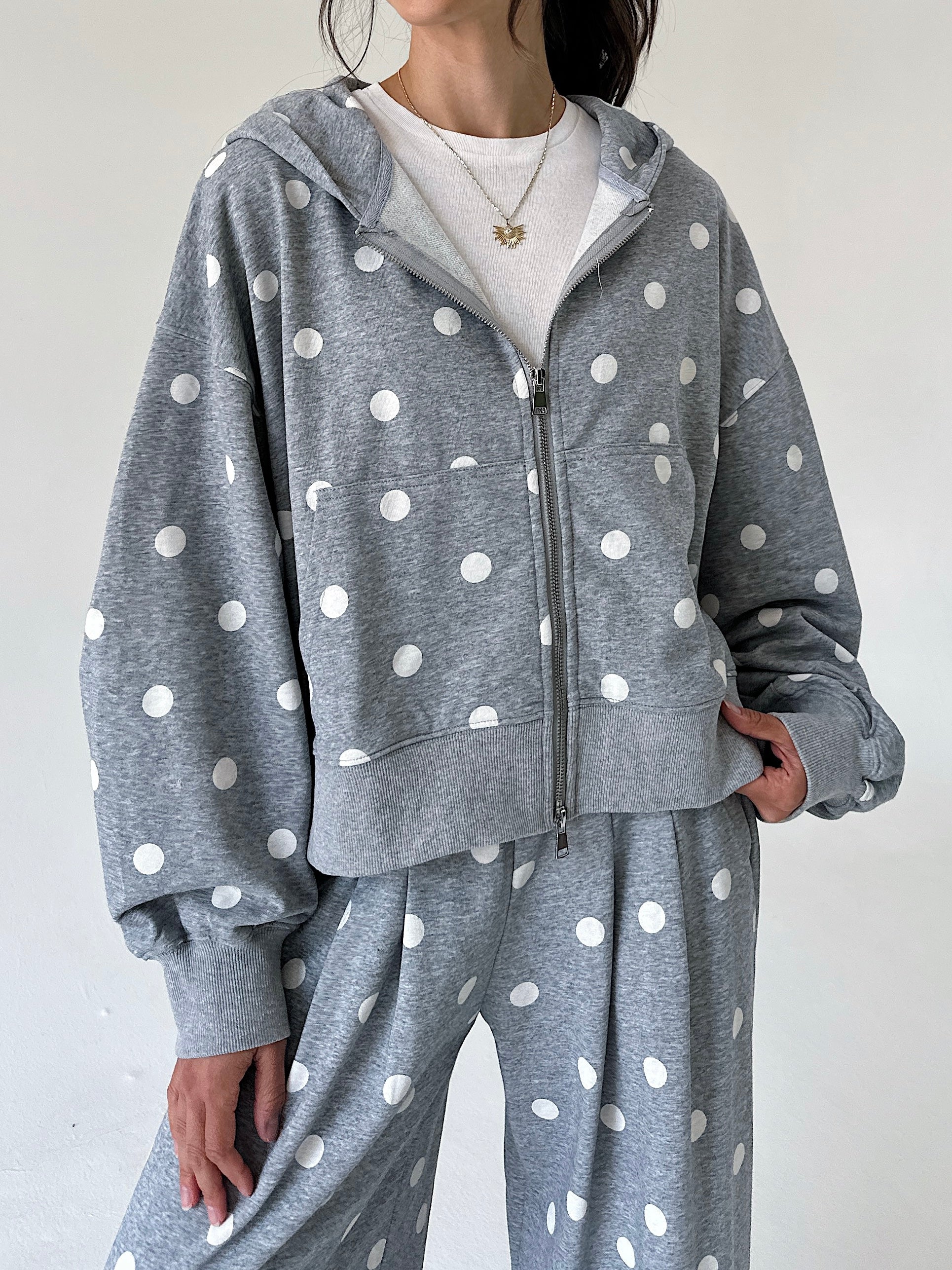 A person wearing a gray zip-up hoodie and matching sweatpants with white polka dots, styled over a white crewneck shirt against a plain light background.