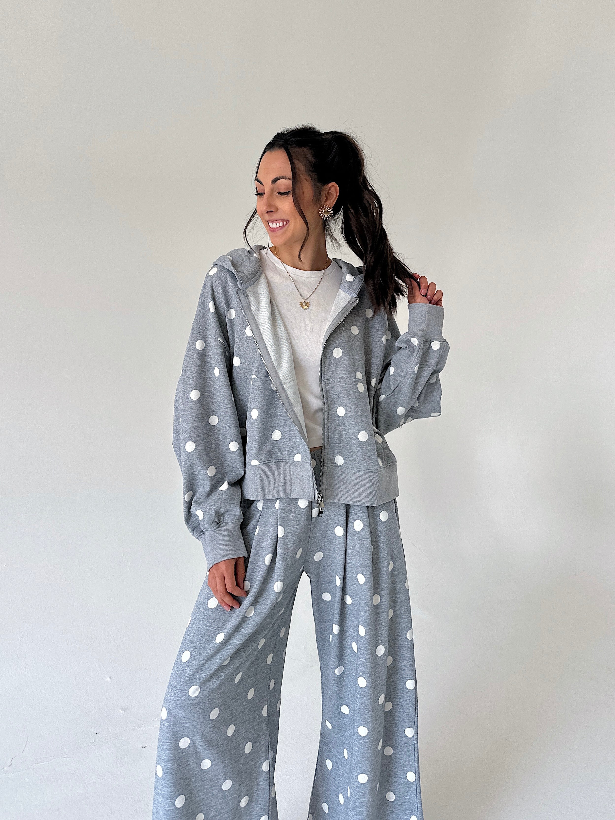 A woman modeling a matching gray and white polka dot lounge set, featuring a zip-up hoodie and wide-leg pants, over a white t-shirt, against a plain off-white background.