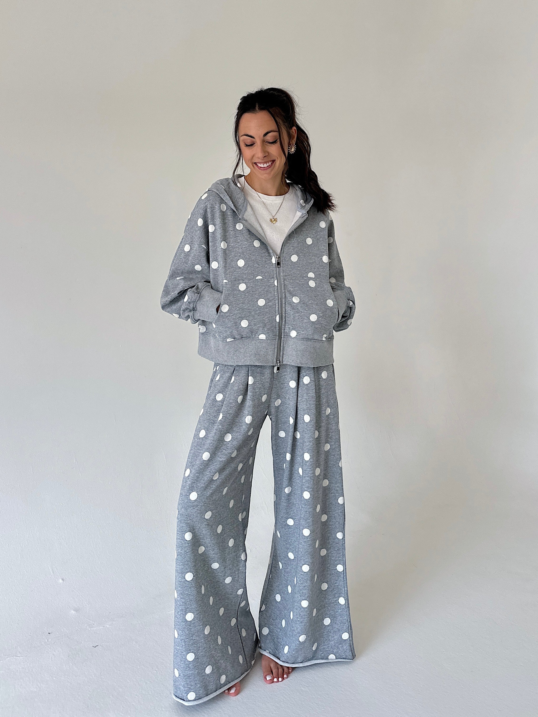 A woman modeling a matching gray and white polka dot loungewear set, consisting of a zip-up hoodie and wide-leg pants, against a plain light gray studio background.
