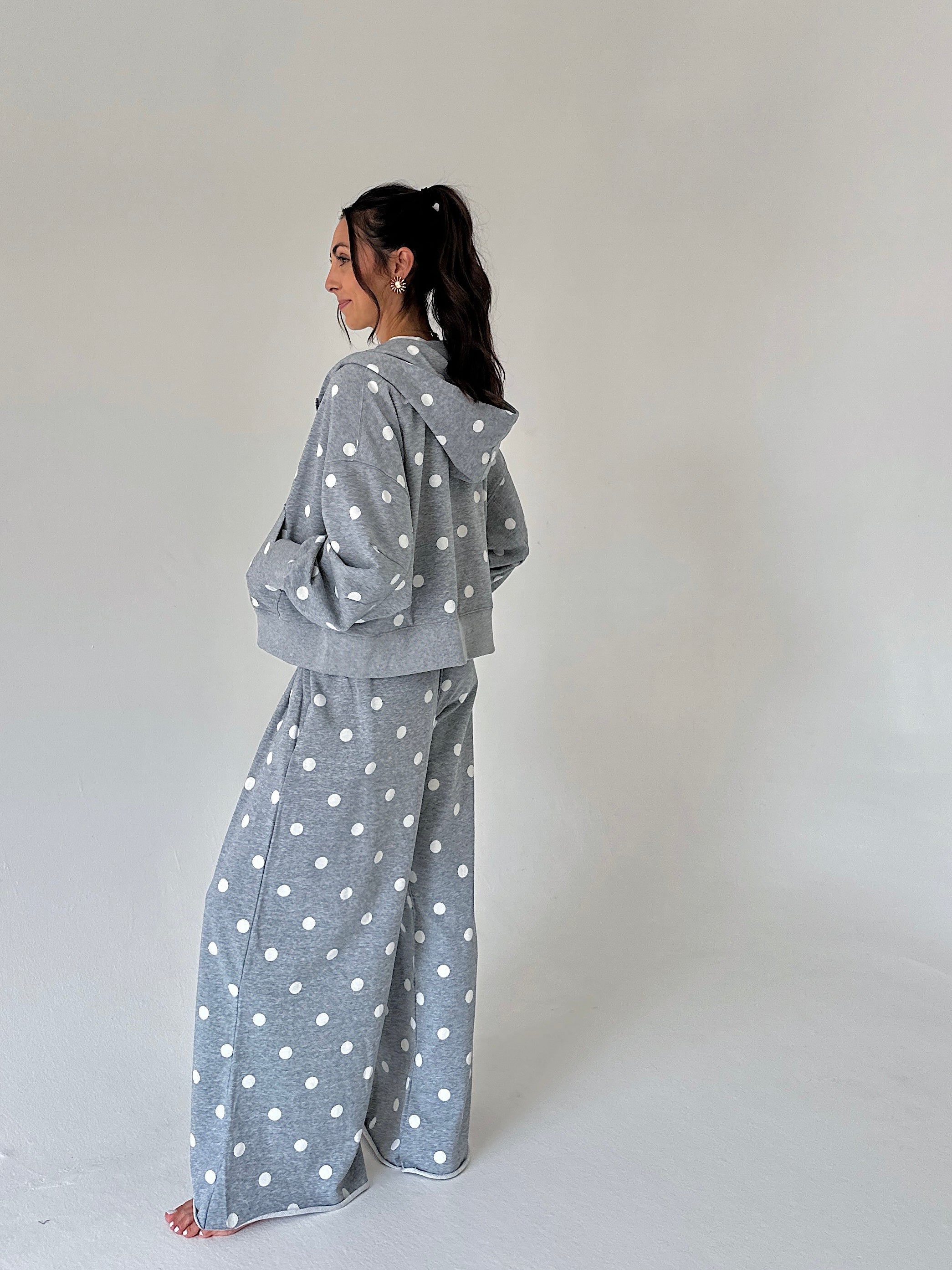 A woman stands in profile wearing a matching gray hooded sweatshirt and wide-leg sweatpants set with white polka dots, photographed against a plain light gray studio background.