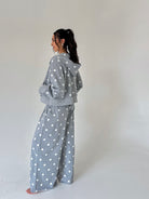 A woman stands in profile wearing a matching gray hooded sweatshirt and wide-leg sweatpants set with white polka dots, photographed against a plain light gray studio background.