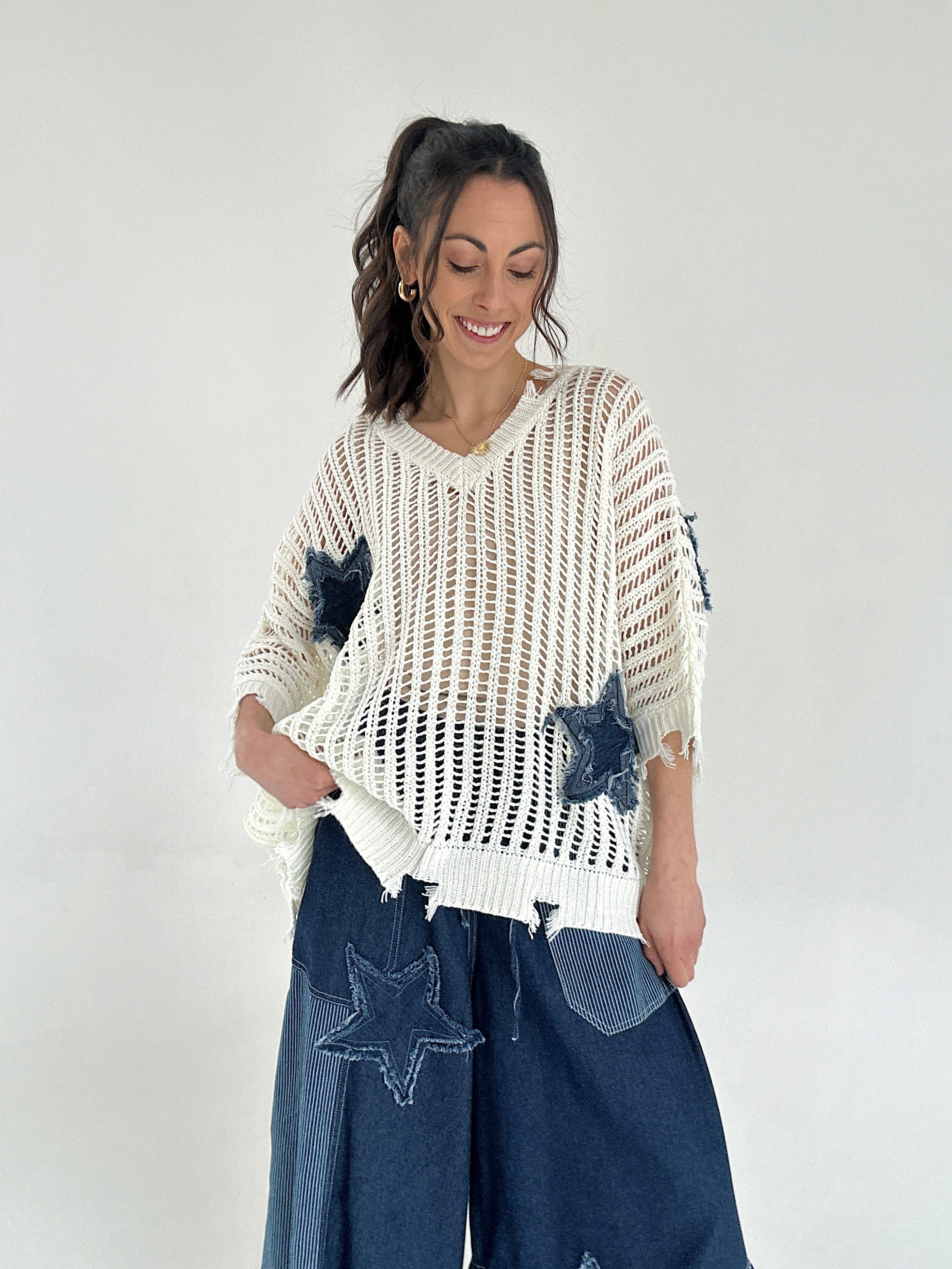 Woman wearing a white knit sweater with star patterns and blue jeans on a plain background