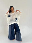 Woman wearing a white knit sweater with star patterns and blue jeans on a plain background