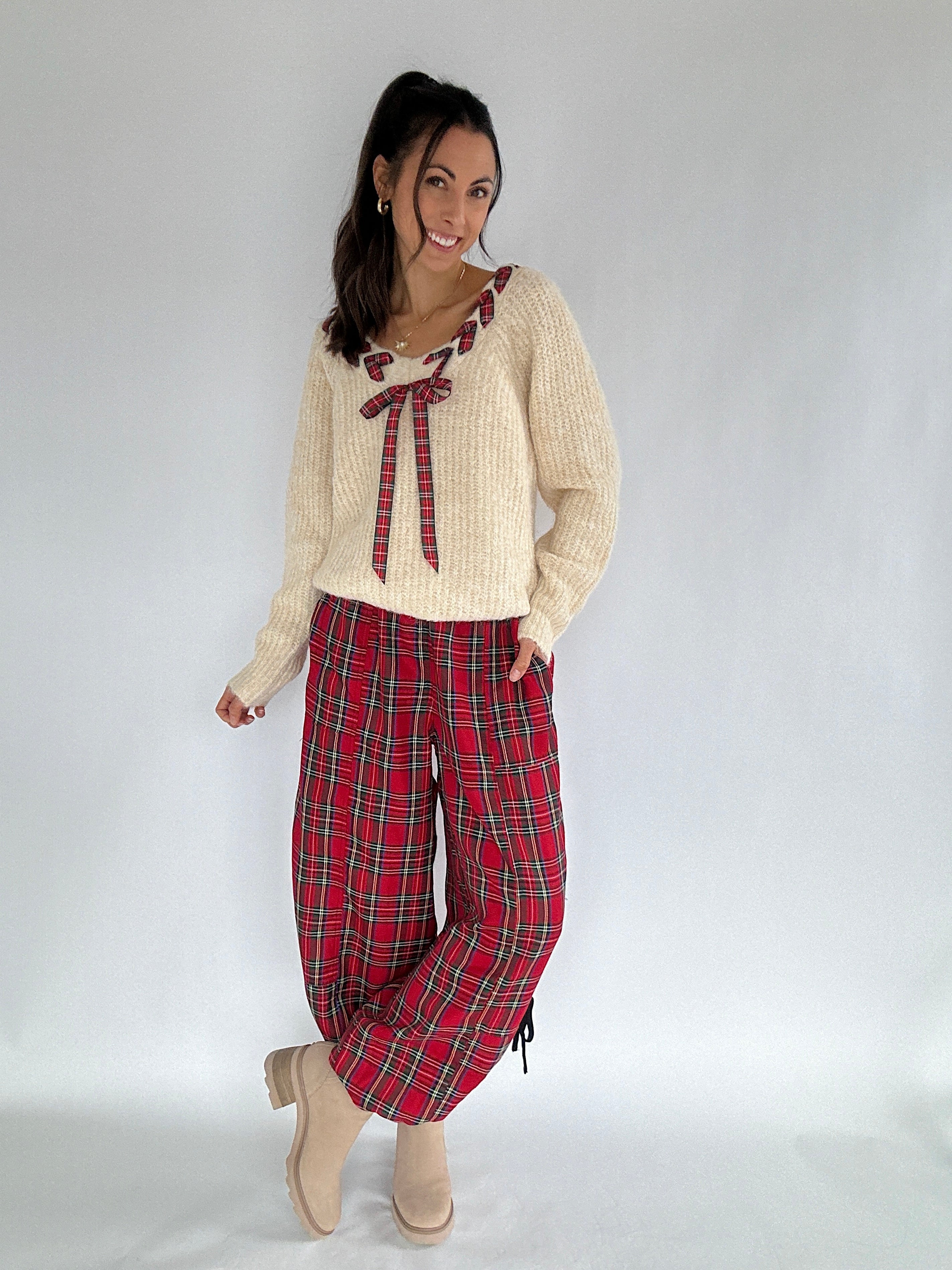 Woman wearing a cream sweater with a red plaid bow and red plaid pants on a white background