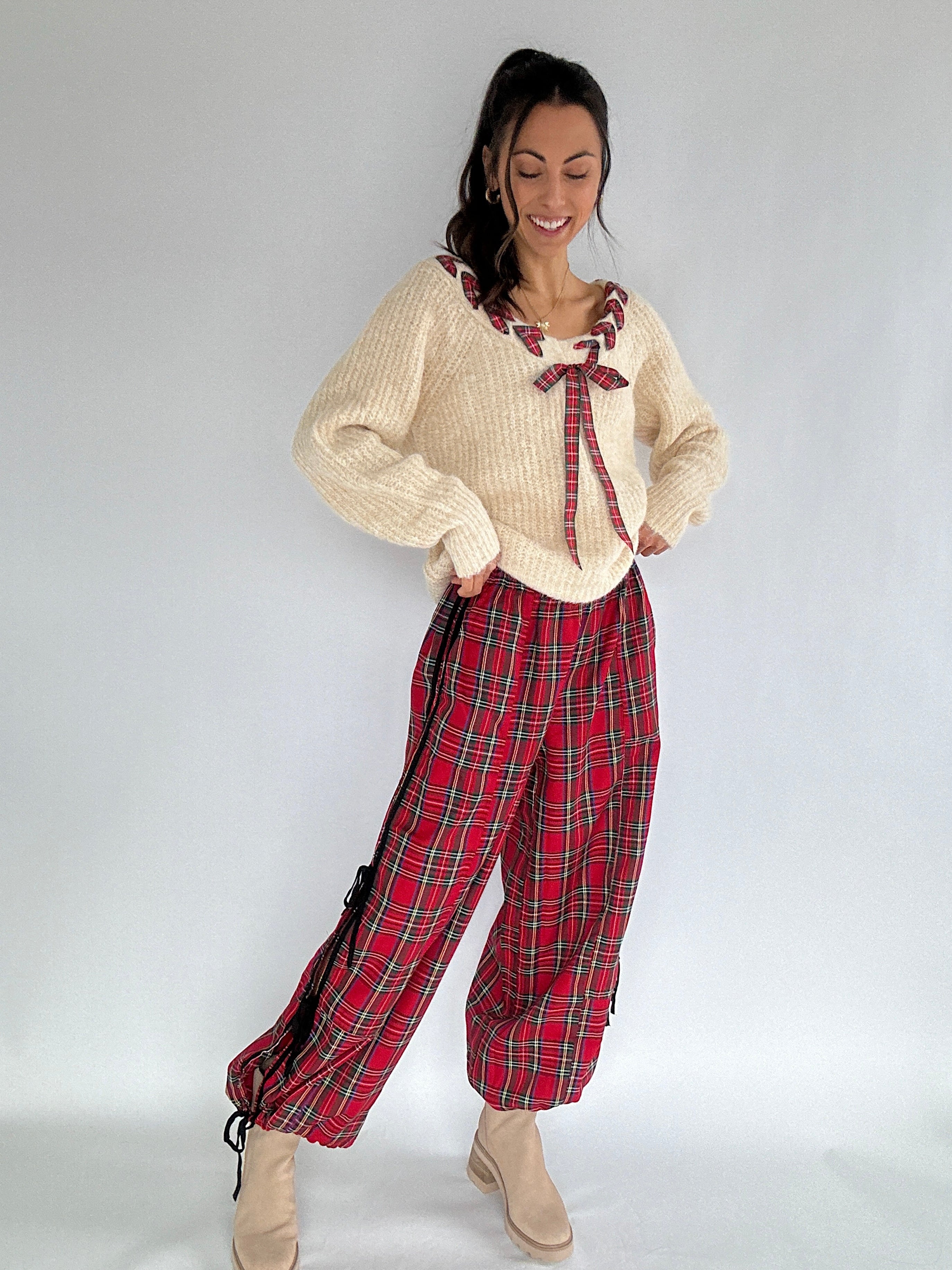 Woman wearing a beige ribbed sweater with a red plaid bow and red plaid pants on a white background