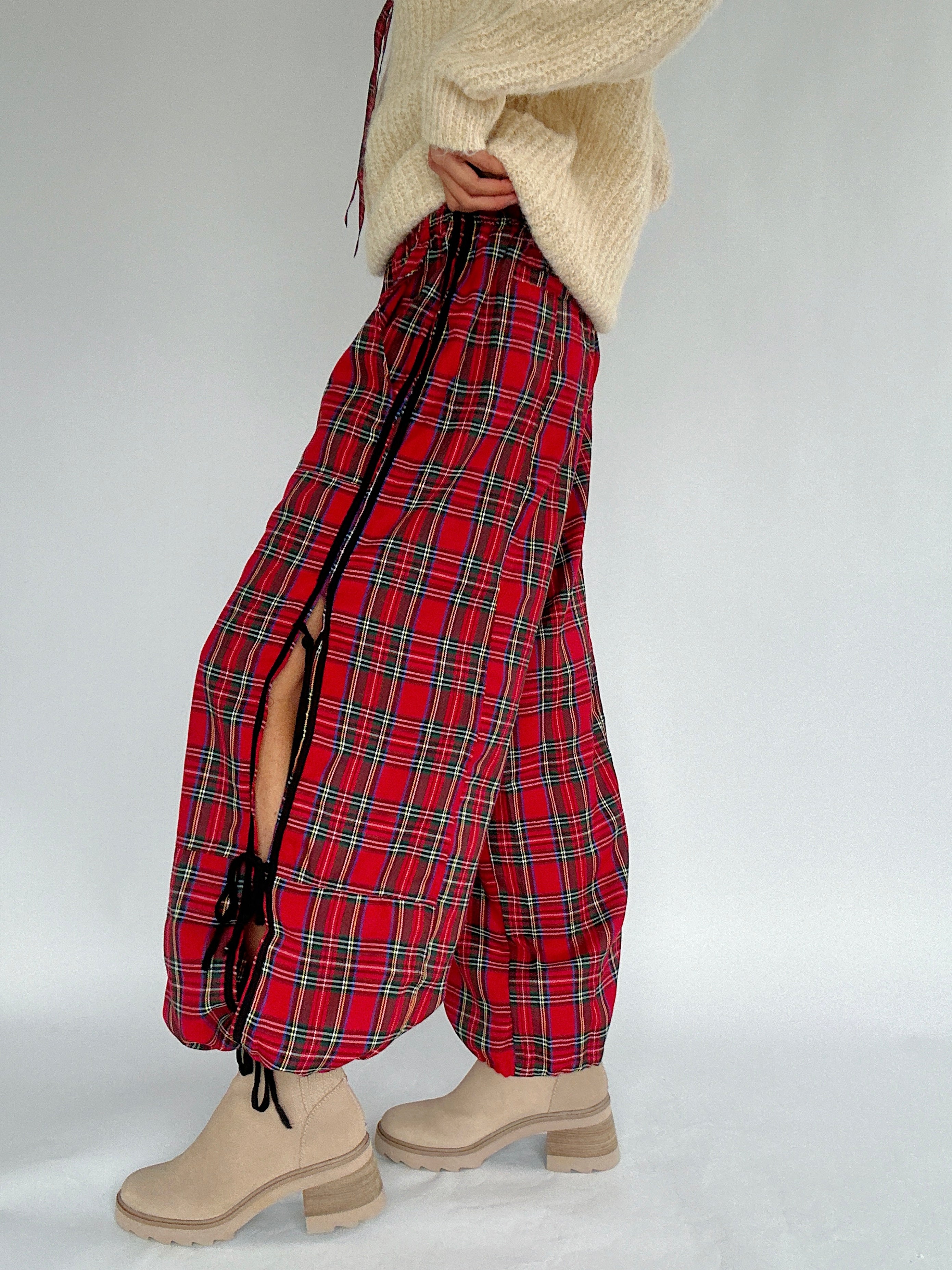 Red plaid pants with side ties worn with a beige sweater and shoes on a light gray background