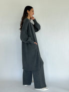 Woman wearing a long dark gray cardigan and matching pants against a white background from side