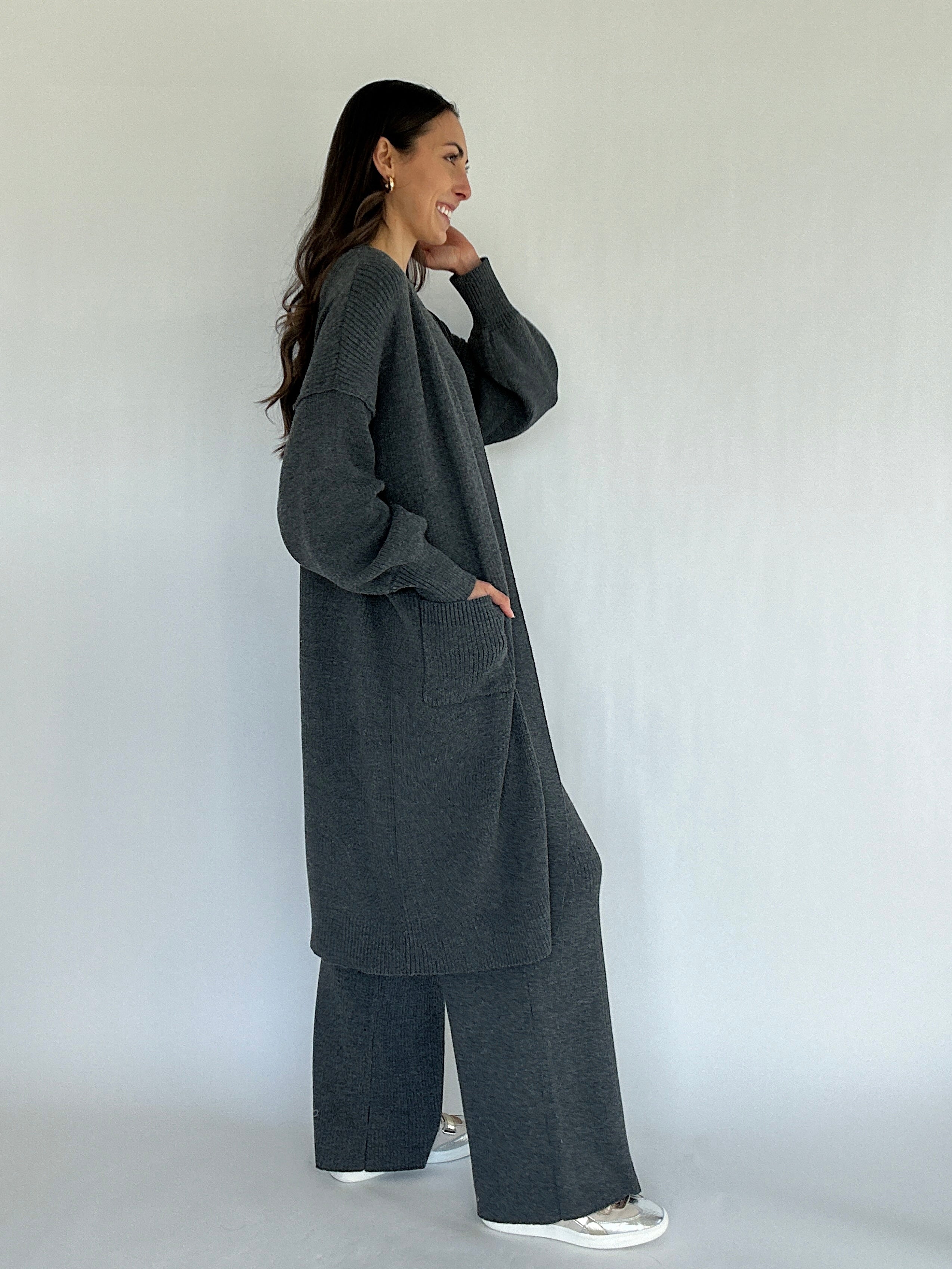 Woman wearing a long dark gray cardigan and matching pants against a white background from side