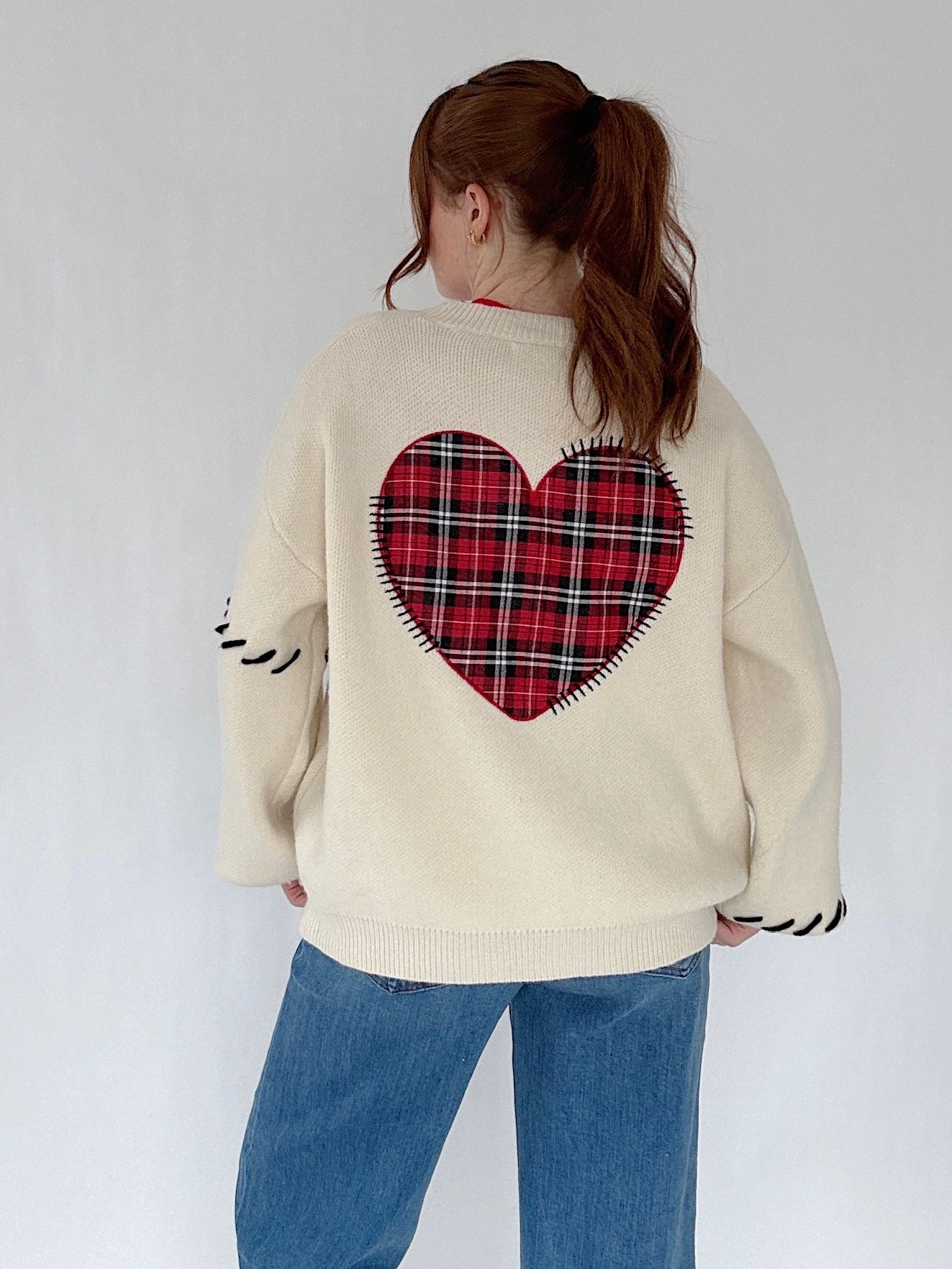 Person wearing a cream sweater with a red plaid heart design on the back, standing against a white background.
