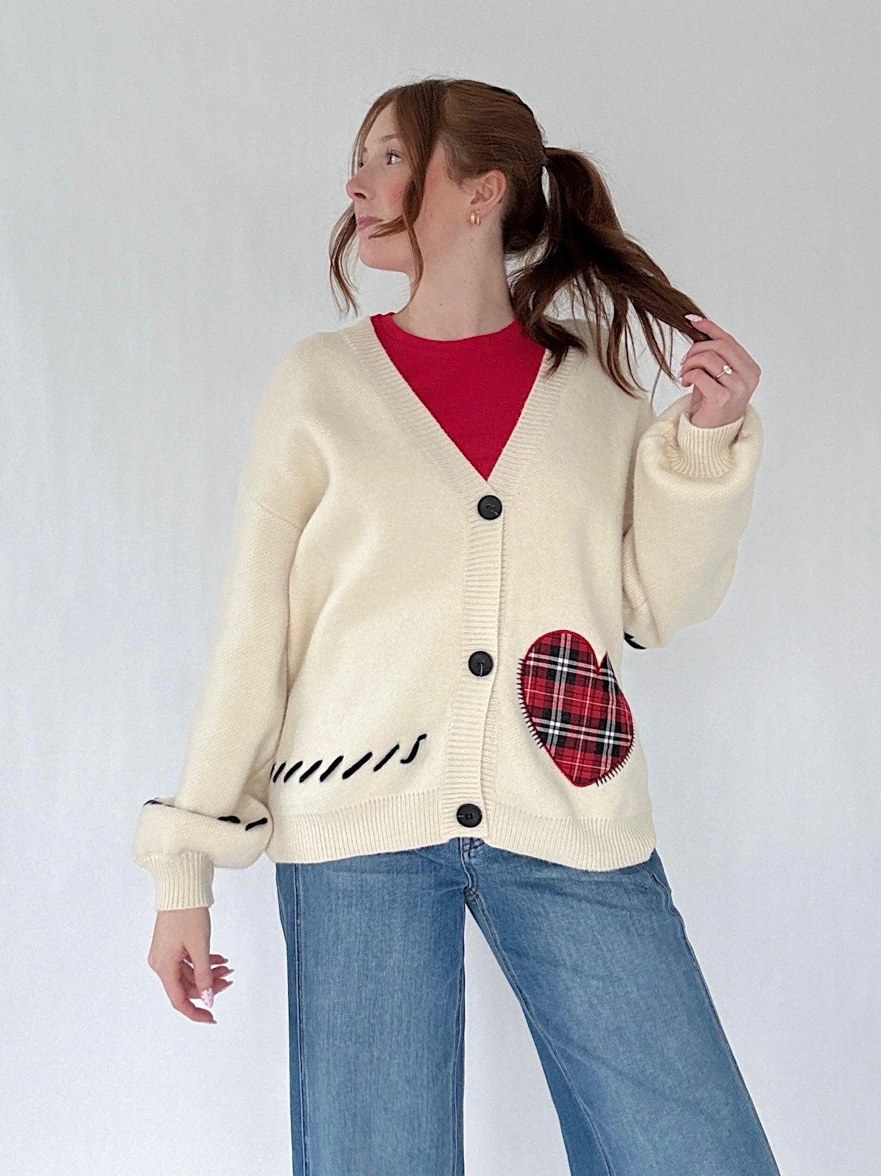 Person wearing a cream cardigan with a red tartan plaid heart patch on a plain background