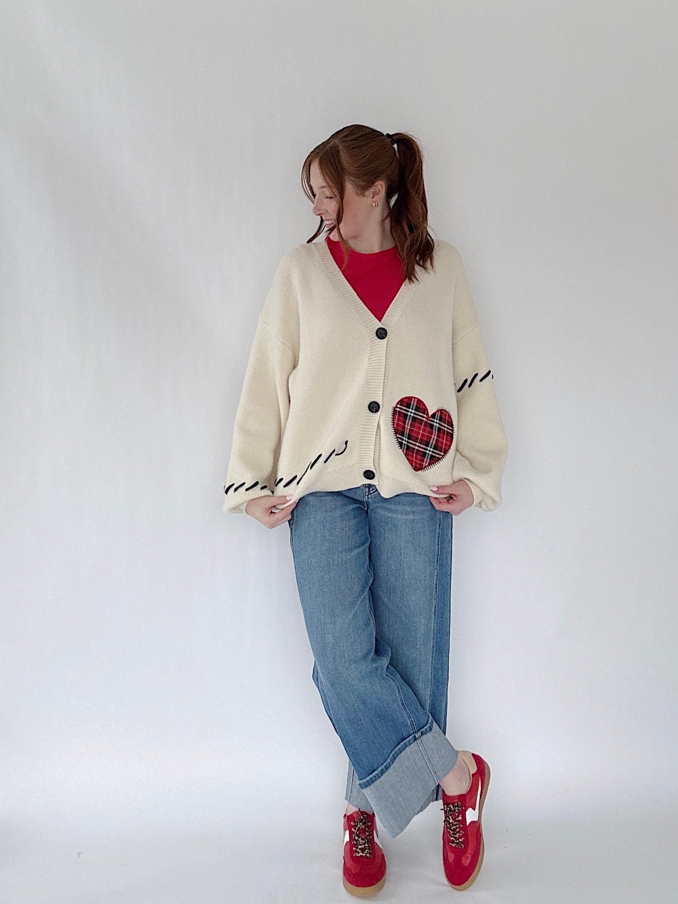 Person wearing a cream cardigan with a heart design, red shirt, blue jeans, and red shoes on a white background