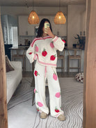 A woman takes a mirror selfie in a cozy room wearing a matching white two-piece set with pink and red strawberry prints. The set includes a long-sleeve top and wide-leg pants, styled with beige slippers.