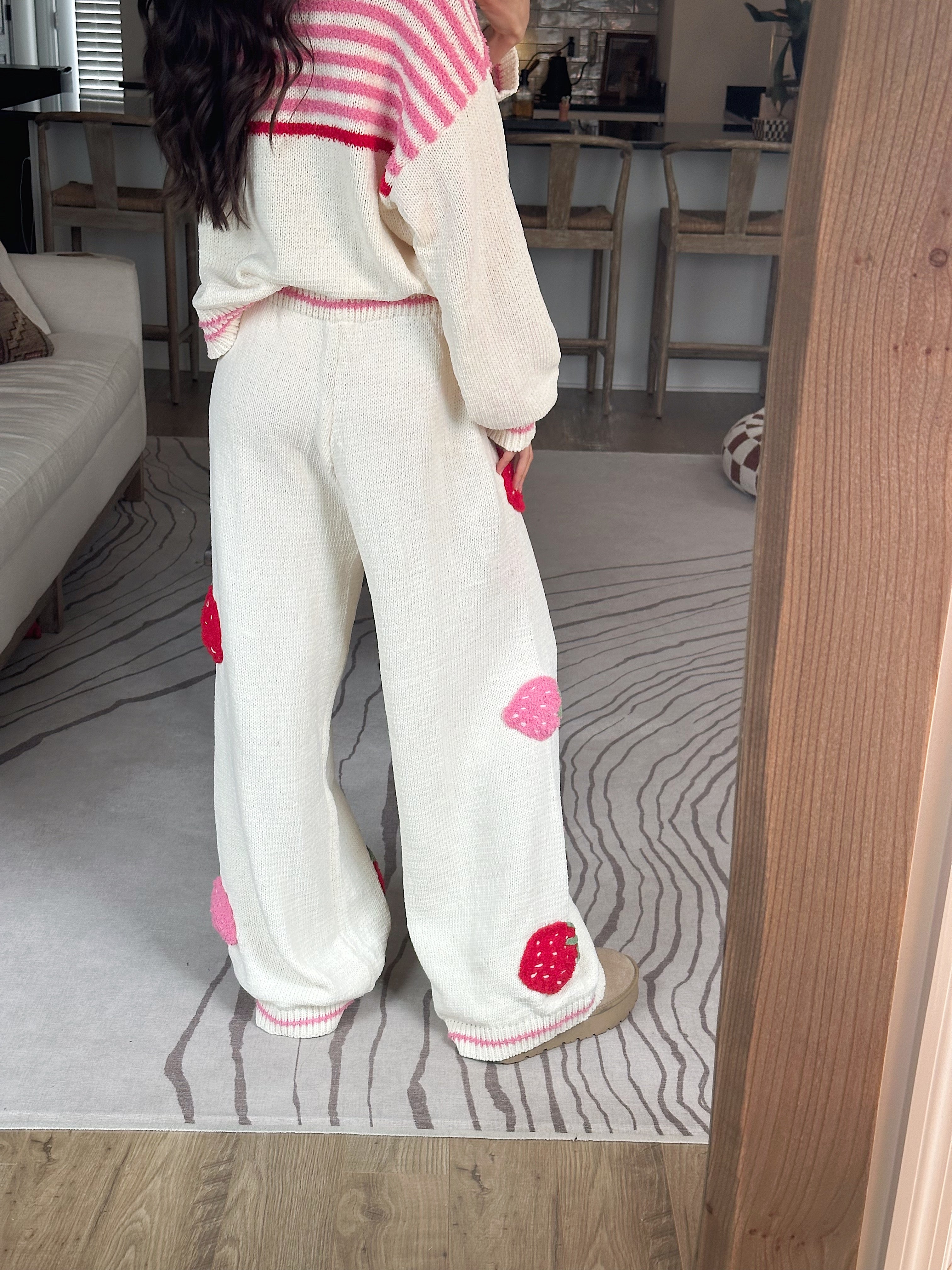 A person wearing a cozy cream knit matching set with pink and red strawberry details showing the backside of the pants, standing on a patterned rug in a home interior.