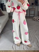 A person wearing a cozy knit matching set with a cream-colored sweater and wide-leg pants, both decorated with 3D strawberry appliques in red and pink, standing on a patterned gray rug in a living room.