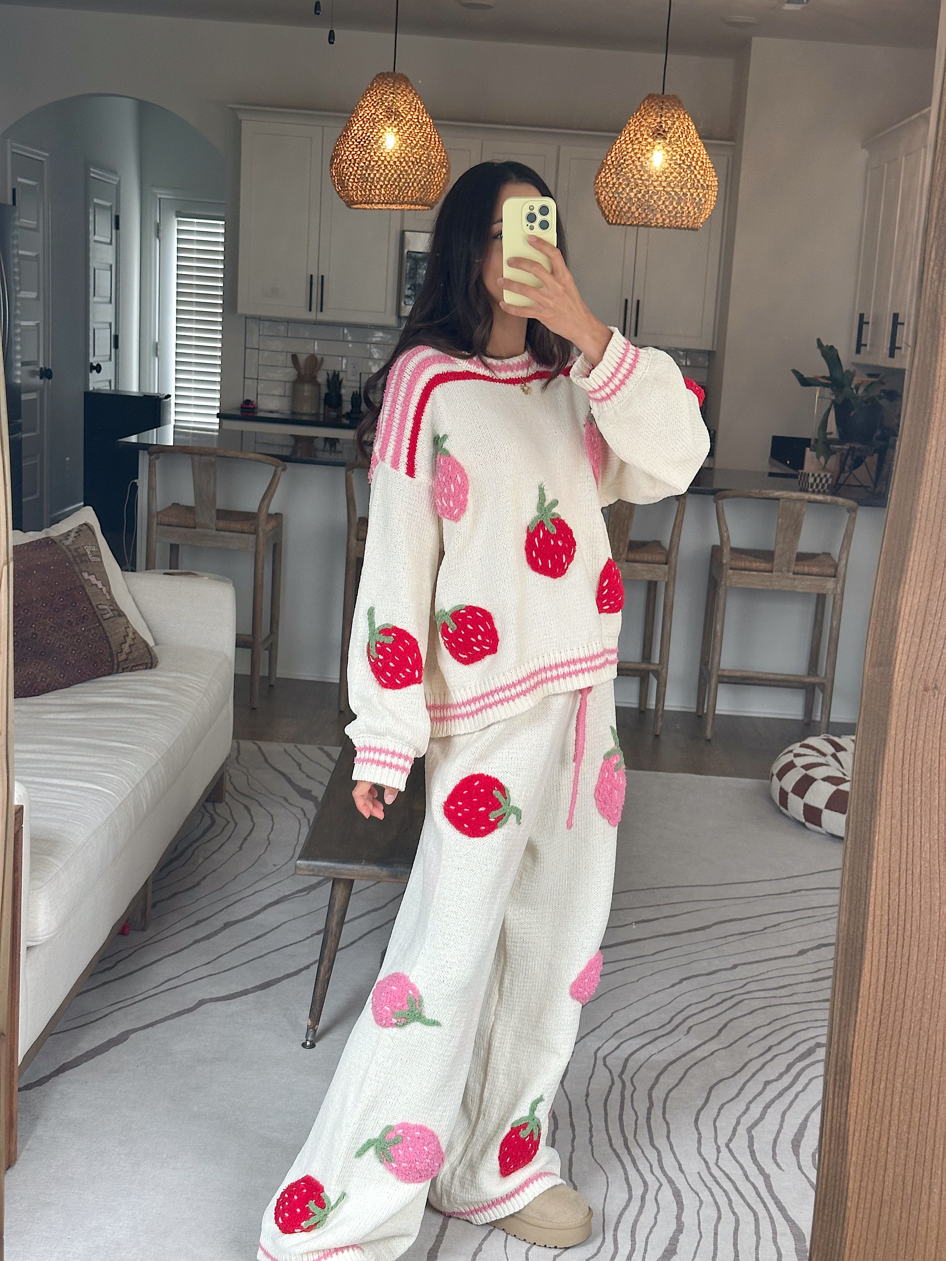 A woman takes a mirror selfie in a cozy living room wearing a matching cream-colored knit lounge set with pink and red strawberry prints. The set includes a long-sleeve sweatshirt and wide-leg pants.