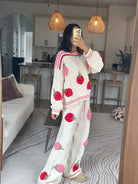 A woman takes a mirror selfie in a cozy living room wearing a matching cream-colored knit lounge set with pink and red strawberry prints. The set includes a long-sleeve sweatshirt and wide-leg pants.