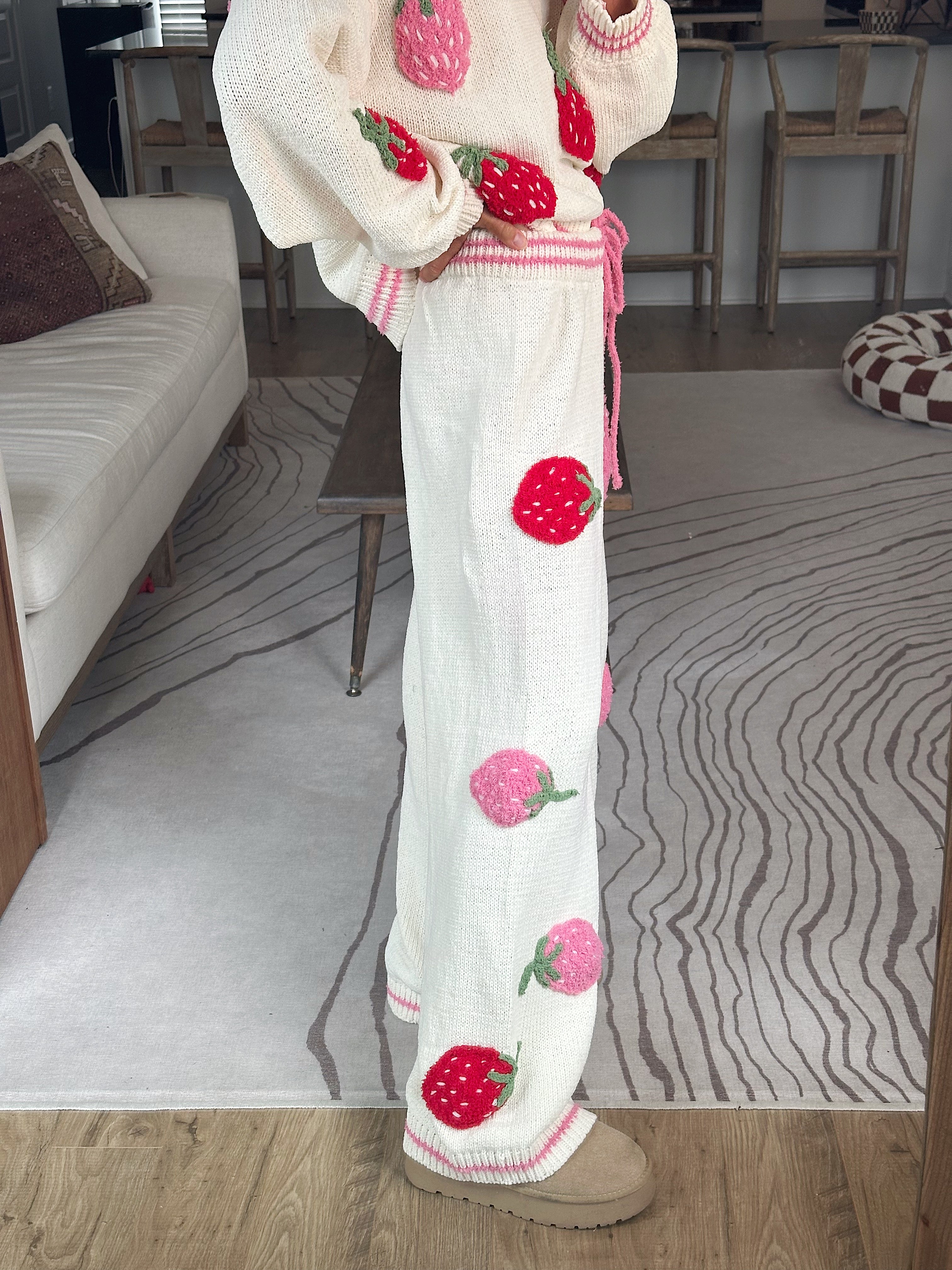 A person wearing a cream knit sweater and matching wide-leg pants with large strawberry appliqués, standing in a living room with a patterned rug and sofa visible.