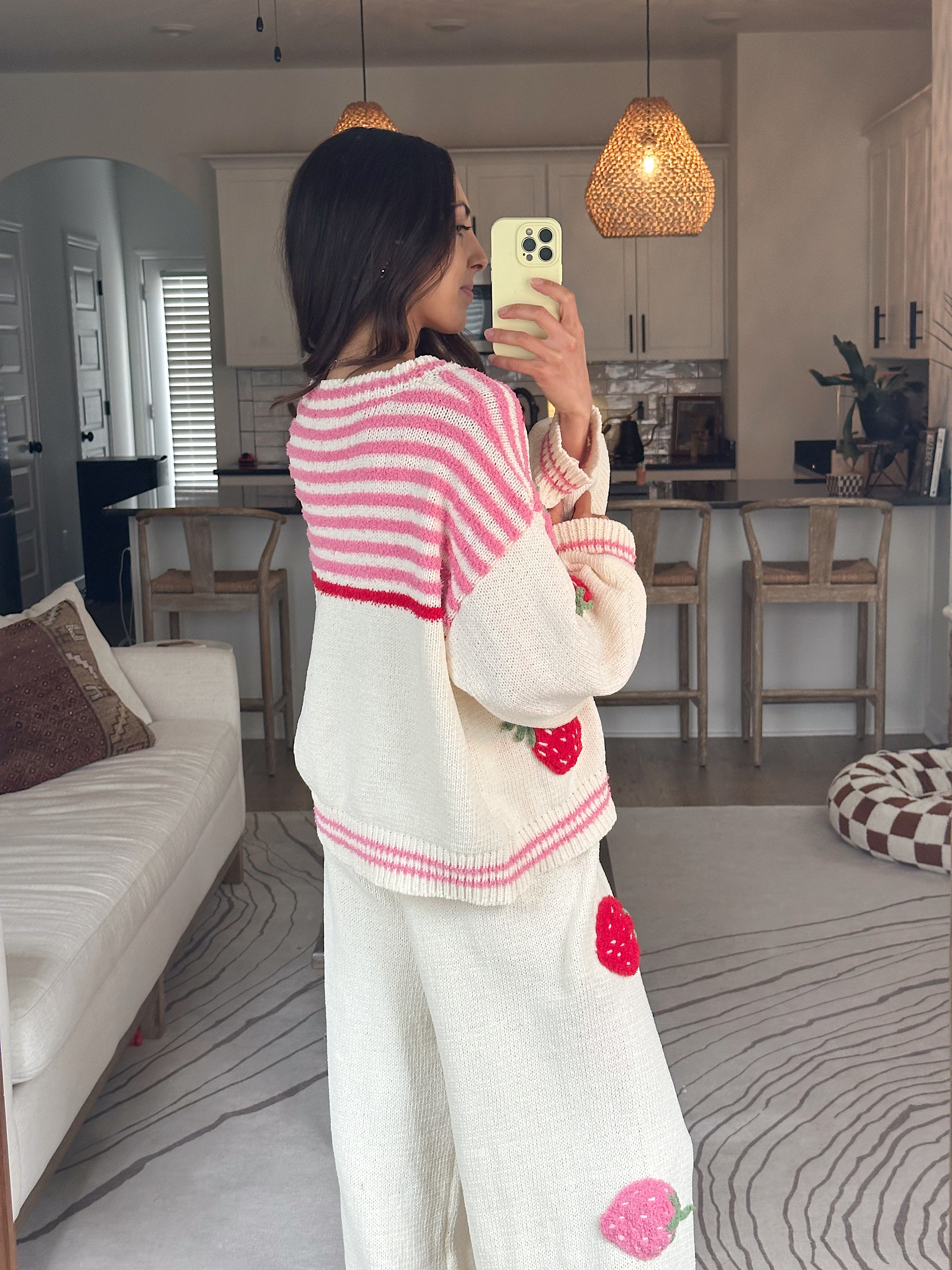 A woman takes a mirror selfie in a cozy living room wearing a cream knit cardigan and matching pants set with pink stripes and embroidered red strawberries.