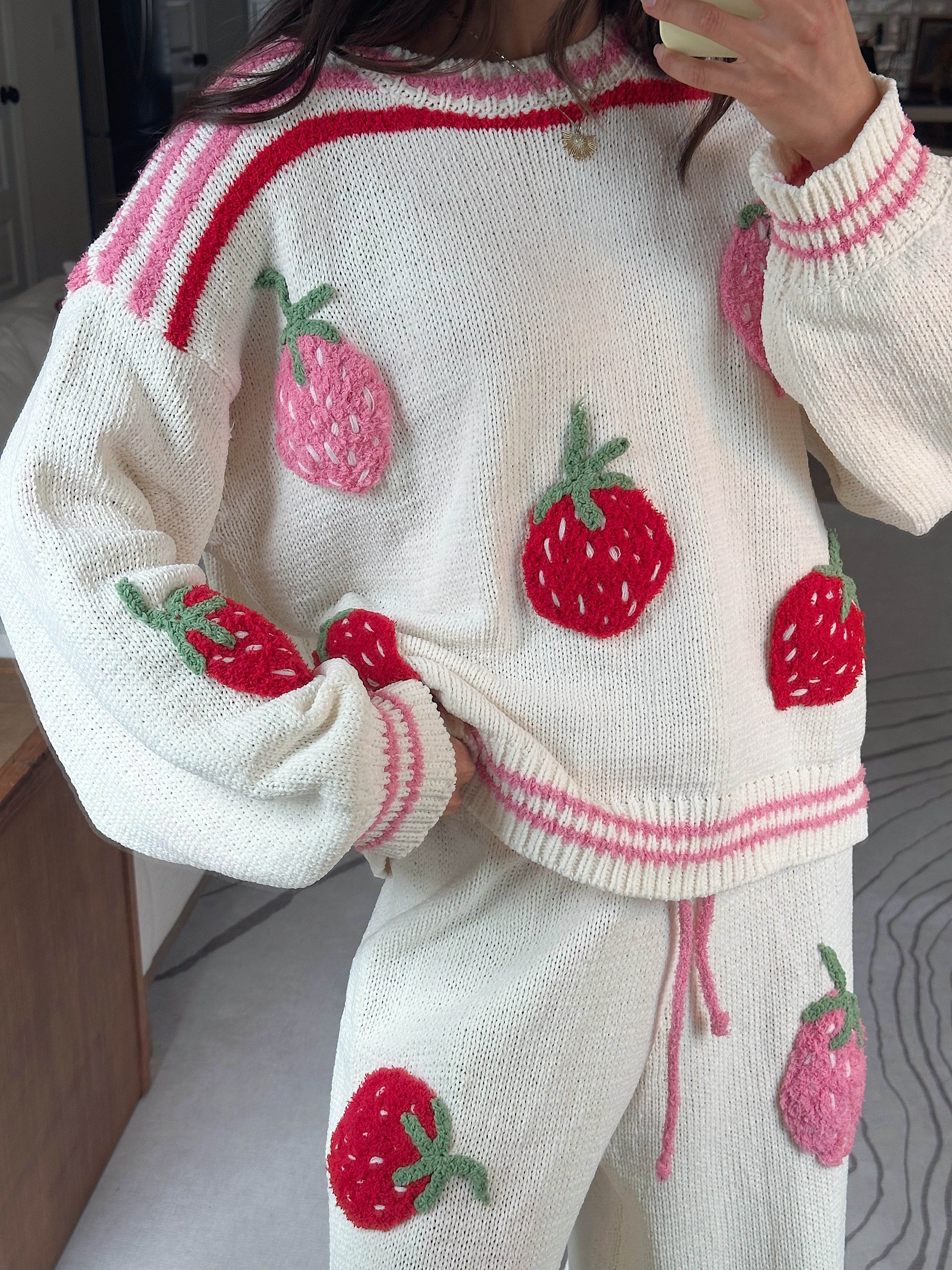 A person wearing a cream knit sweater and matching pants set with pink and red striped trim and large red strawberry appliqués, photographed indoors in a mirror selfie style.
