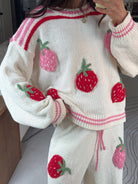 A person wearing a cream knit sweater and matching pants set with pink and red striped trim and large red strawberry appliqués, photographed indoors in a mirror selfie style.