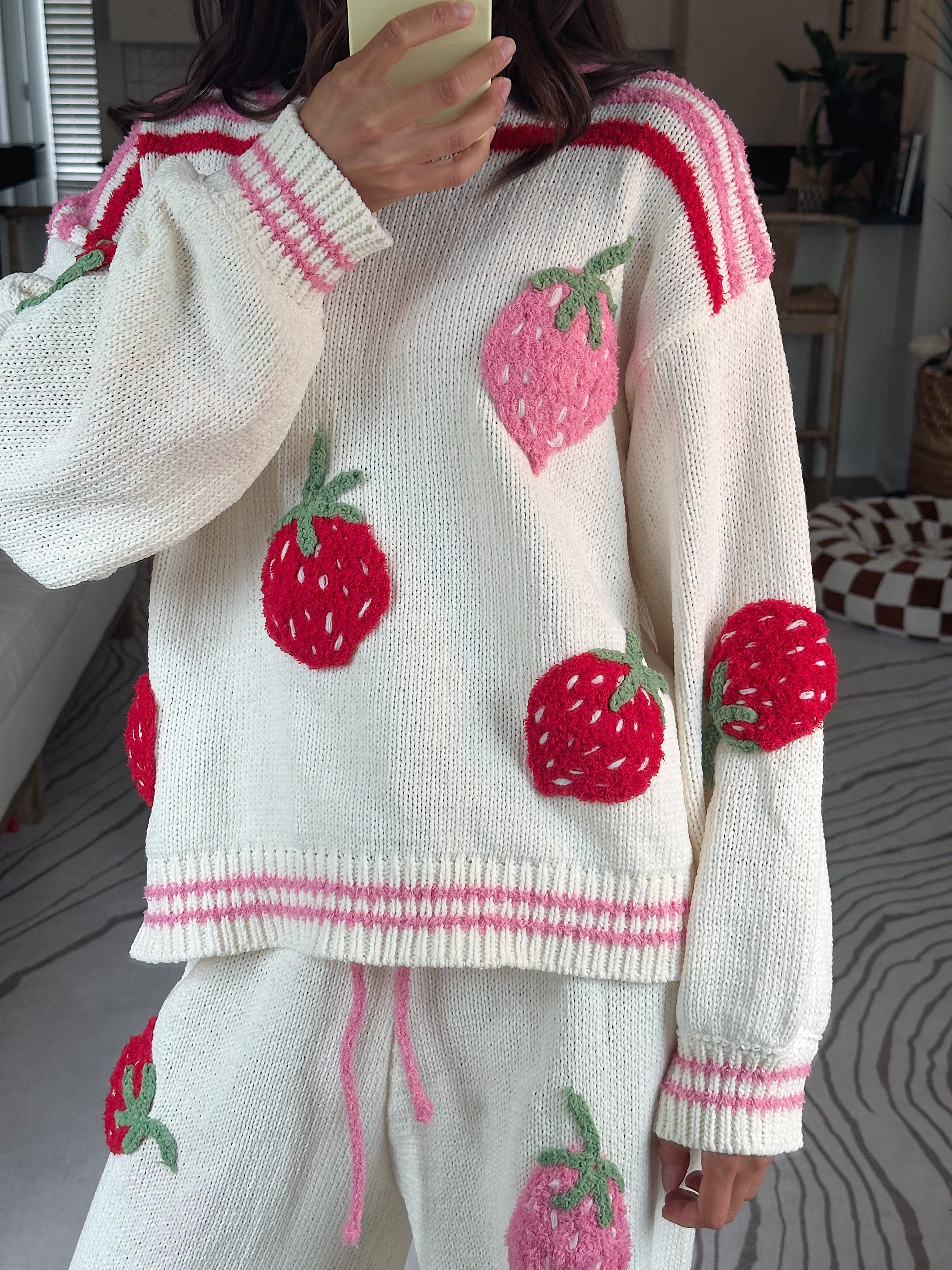 A person wearing a cream knit sweater and matching pants with large red and pink strawberry appliqués, photographed indoors in a home setting.