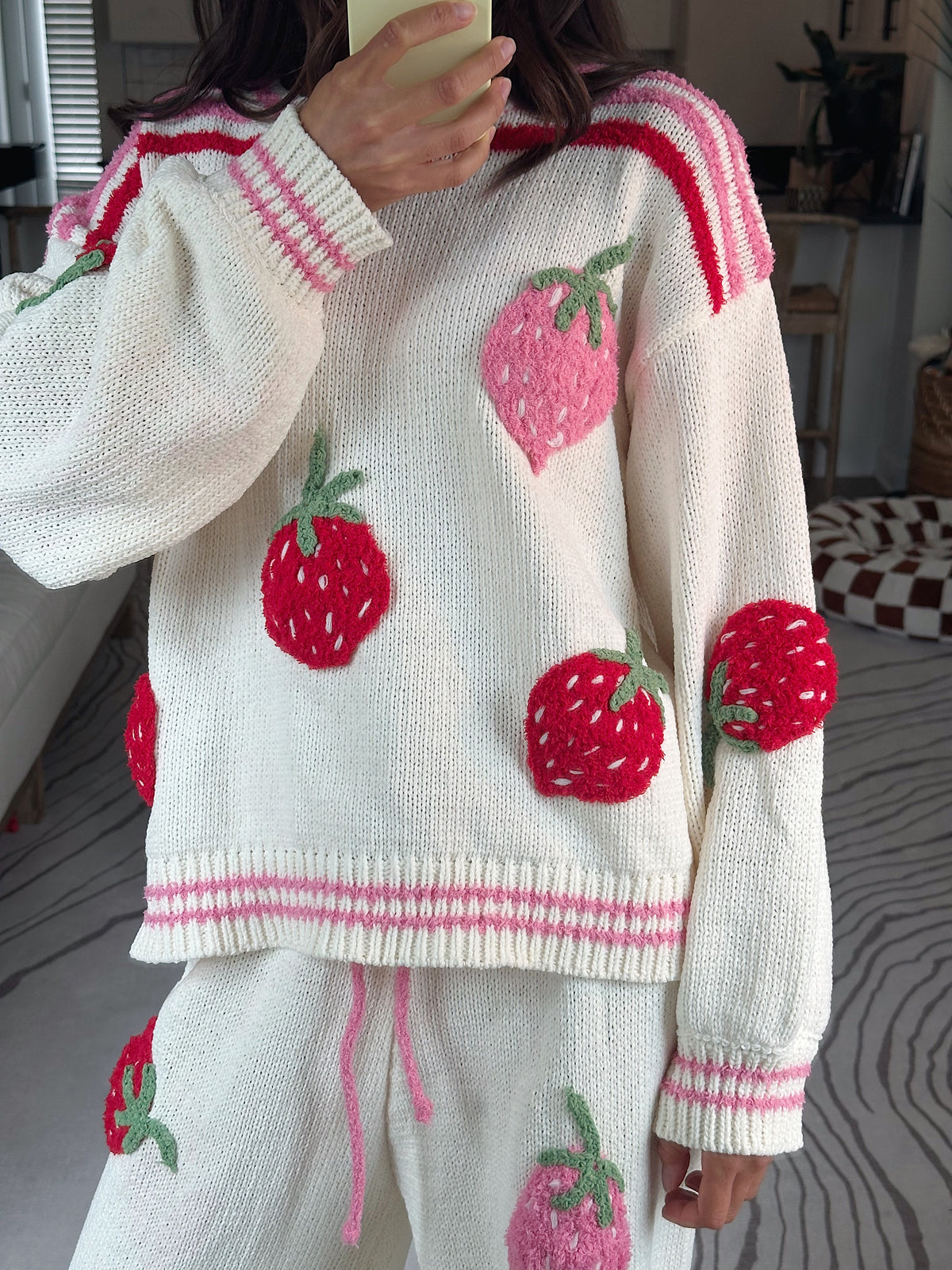 A person wearing a cream knit sweater and matching pants with large red and pink strawberry appliqués, photographed indoors in a home setting.