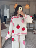 A woman takes a mirror selfie wearing a cozy white knit sweater and matching sweatpants set, both decorated with large red and pink strawberry motifs. The background shows a modern living space with pendant lights, a dining table, and a sofa.