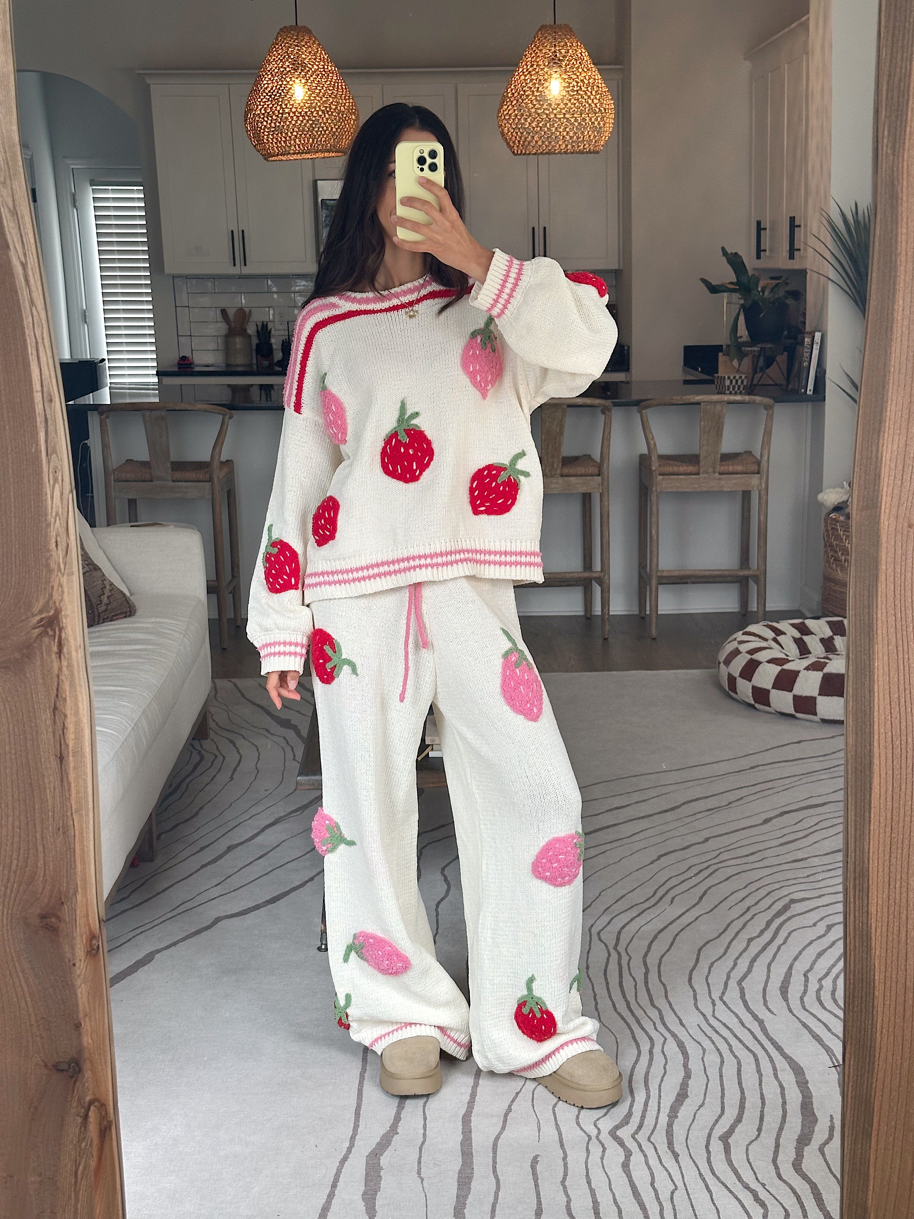 A woman takes a mirror selfie wearing a cozy white knit pajama set featuring a strawberry print and pink striped trim, set in a modern living room with wooden pendant lights and a gray patterned rug.