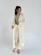Woman wearing a long, light-colored cardigan over a comfy outfit on a plain background