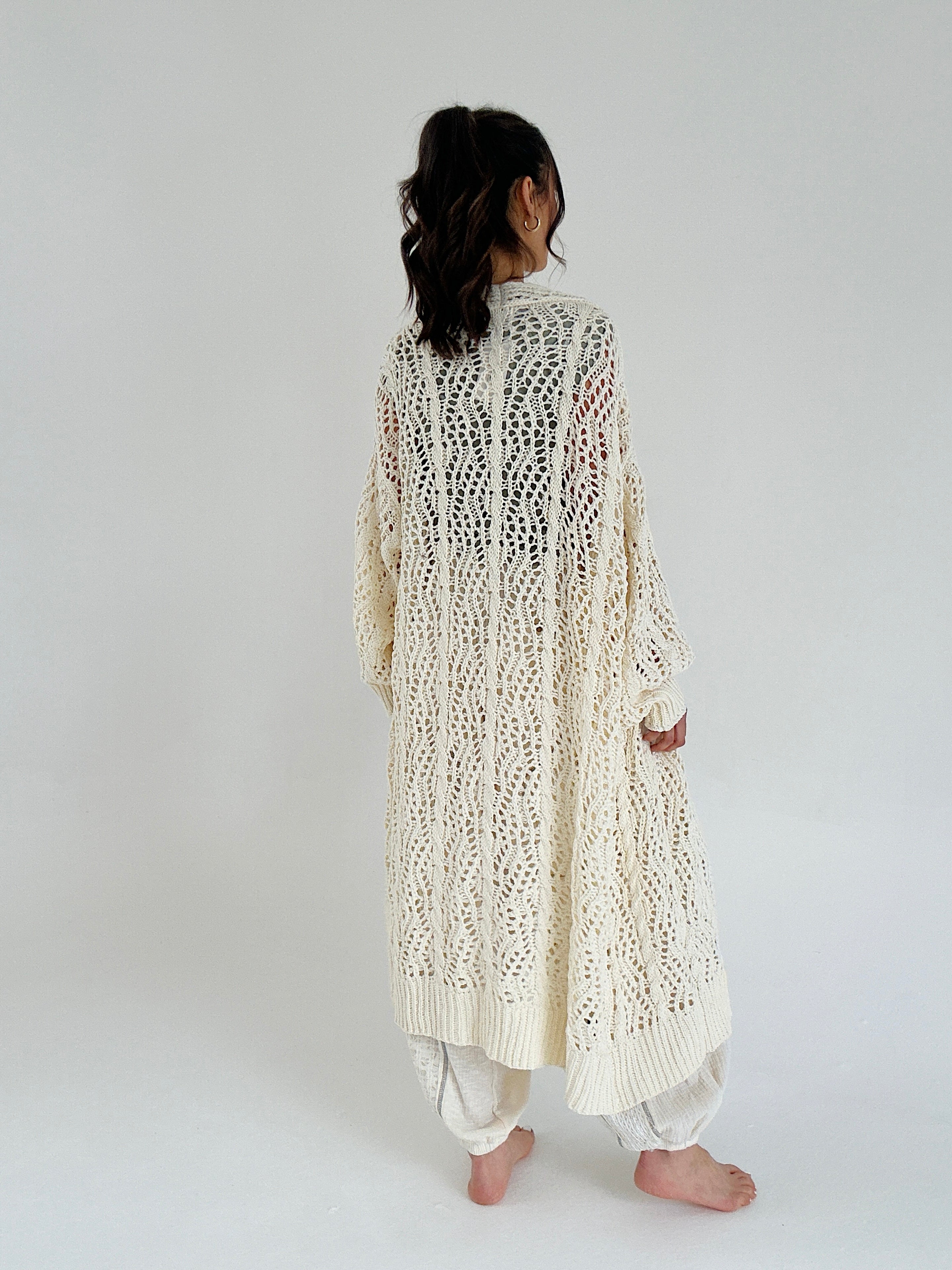 Person wearing a long, cream, knitted cardigan on a plain background showing back