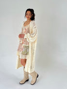 Woman wearing a floral dress and long beige cardigan with white boots on a plain background