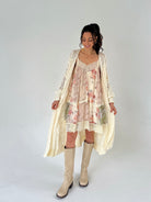 Woman wearing a floral mini dress with a long cream cardigan and knee-high boots on a white background