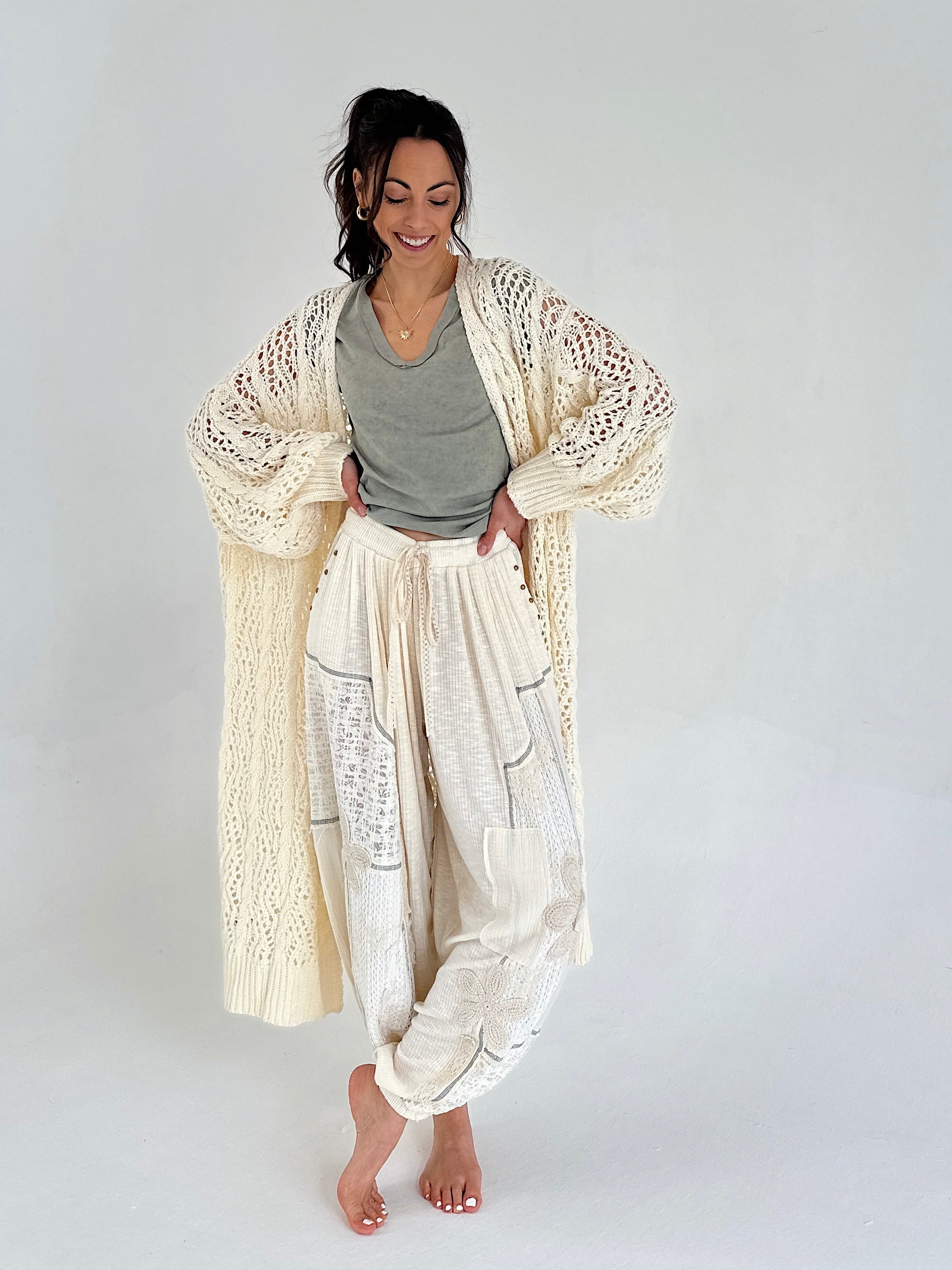 Woman wearing a long, light-colored cardigan over a green/gray top and white pants on a plain background