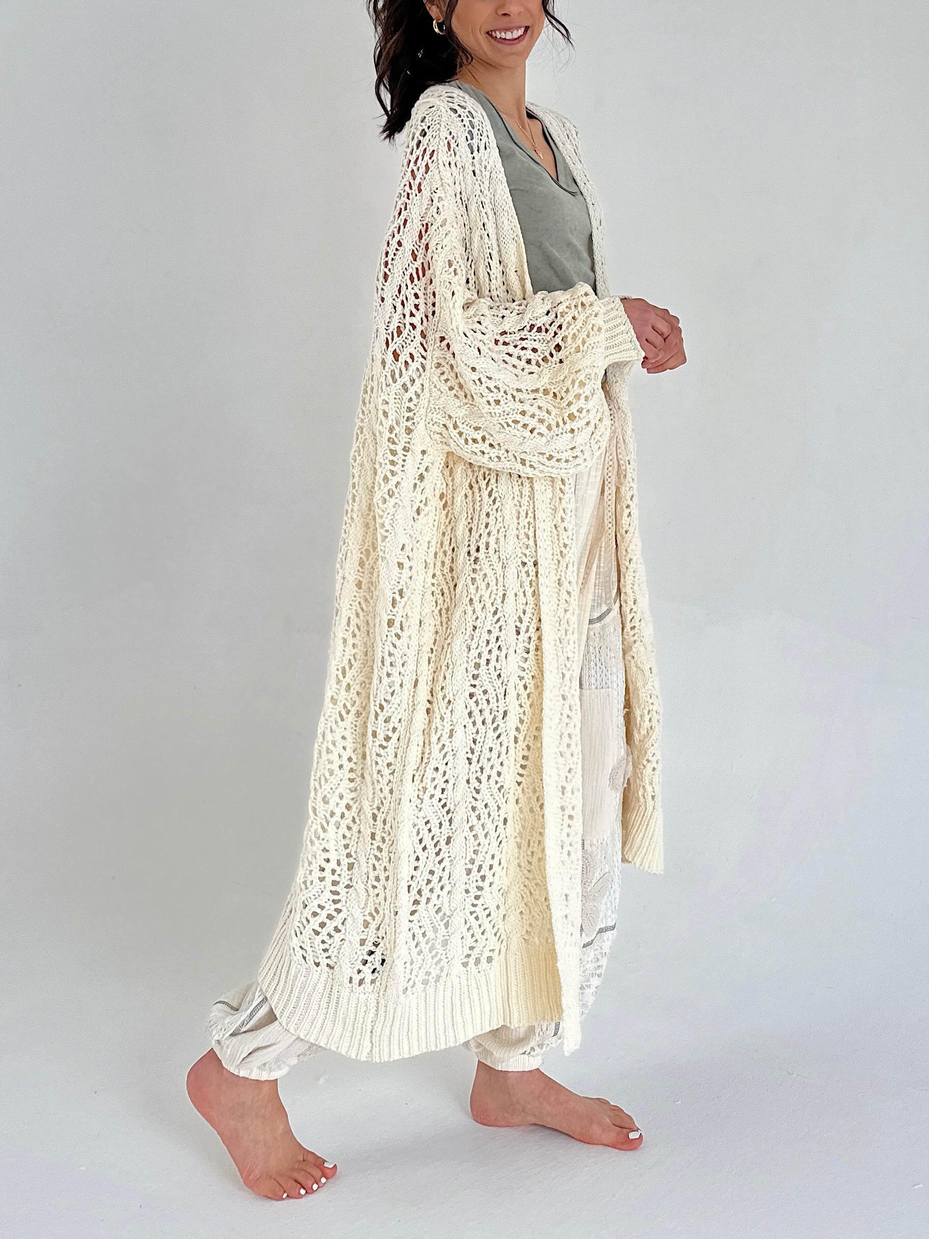 Woman wearing a long, light-colored crochet cardigan on a plain background