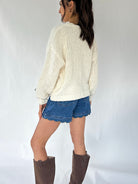 A woman shown from the back wearing a cream cable-knit sweater, blue denim shorts with a scalloped edge, and brown knee-high boots, standing against a white studio backdrop.