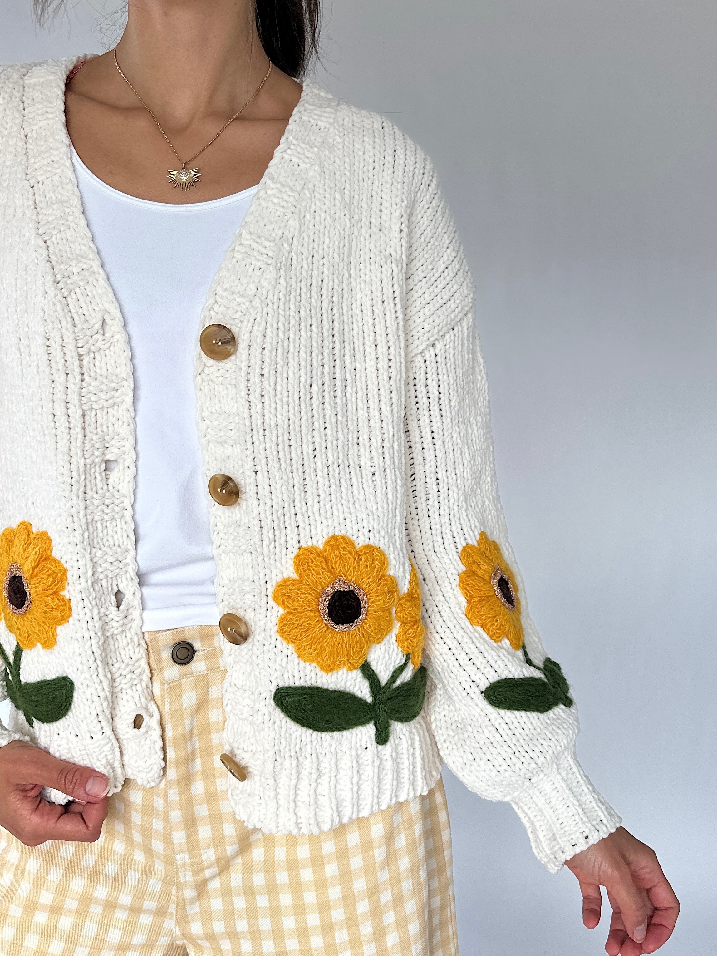 A person wearing a cream knit cardigan with large yellow sunflower patches on the front and sleeves, over a white top and yellow-and-white gingham pants, against a plain light gray background.