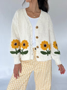 A woman wearing a cream-colored ribbed knit cardigan with large yellow and green sunflower appliqués on the front, paired with a white top and yellow-and-white gingham pants.