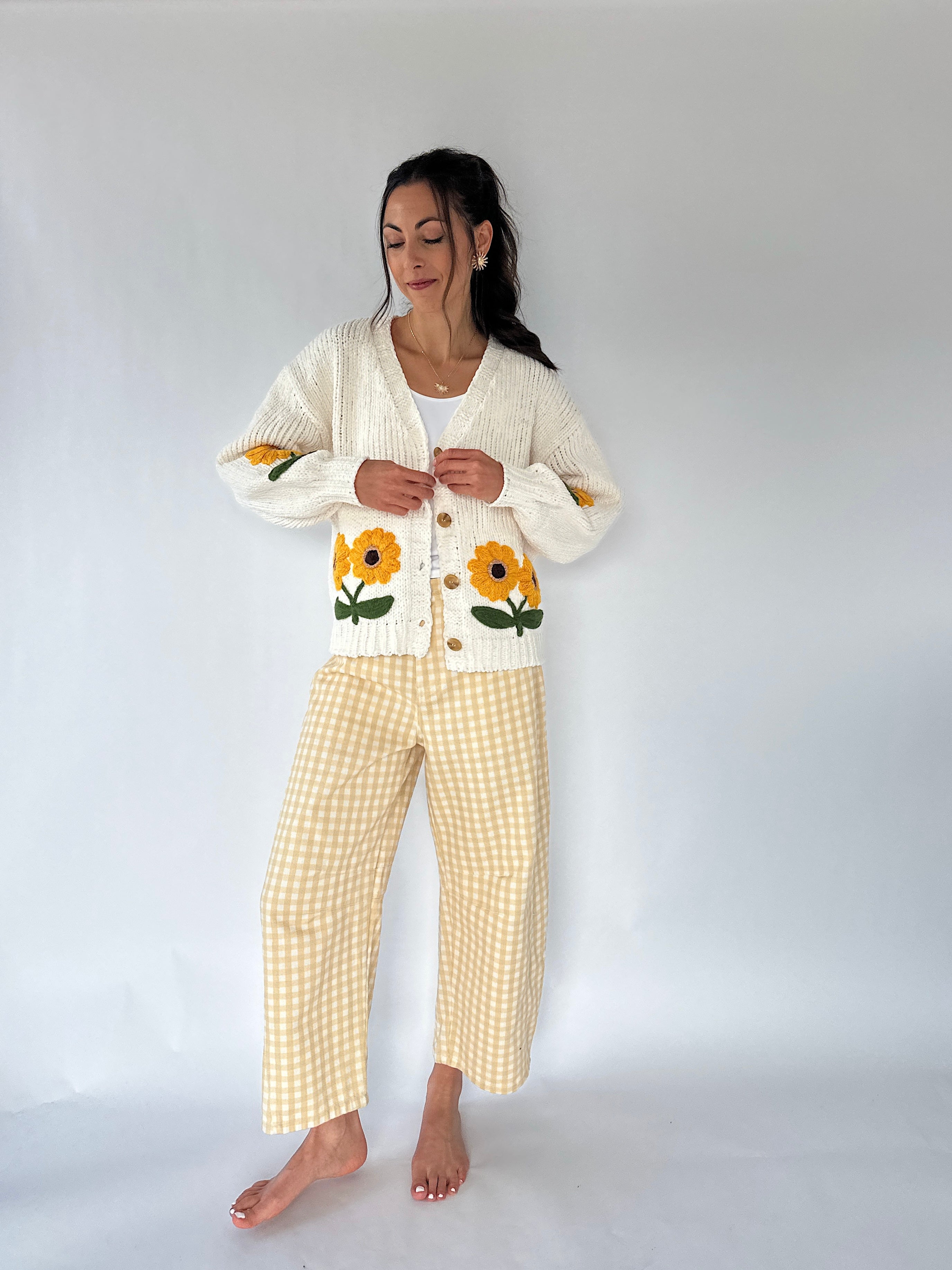 A woman stands barefoot against a plain light gray studio background wearing a cream knit cardigan with yellow sunflower embroidery and yellow-and-white gingham cropped pants.