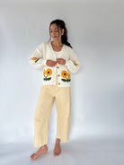 A woman stands barefoot against a plain light gray studio background wearing a cream knit cardigan with yellow sunflower embroidery and yellow-and-white gingham cropped pants.