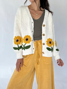 A woman wearing a cream knit open cardigan with large yellow sunflower and green leaf embroidery on the front, paired with a dark olive tank top and mustard yellow wide-leg pants, against a plain light gray studio background.