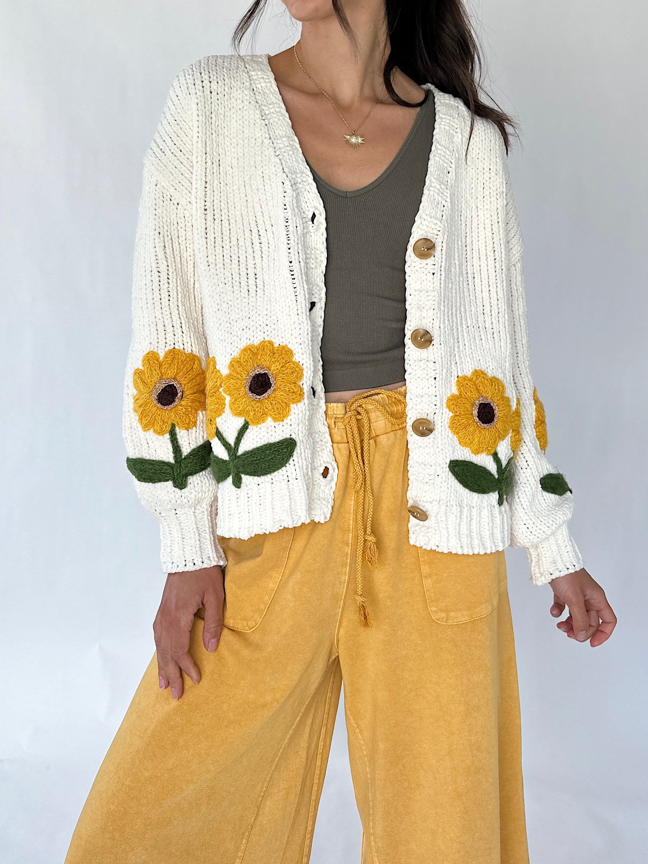 A woman wearing a cream knit open cardigan with large yellow sunflower and green leaf embroidery on the front, paired with a dark olive tank top and mustard yellow wide-leg pants, against a plain light gray studio background.