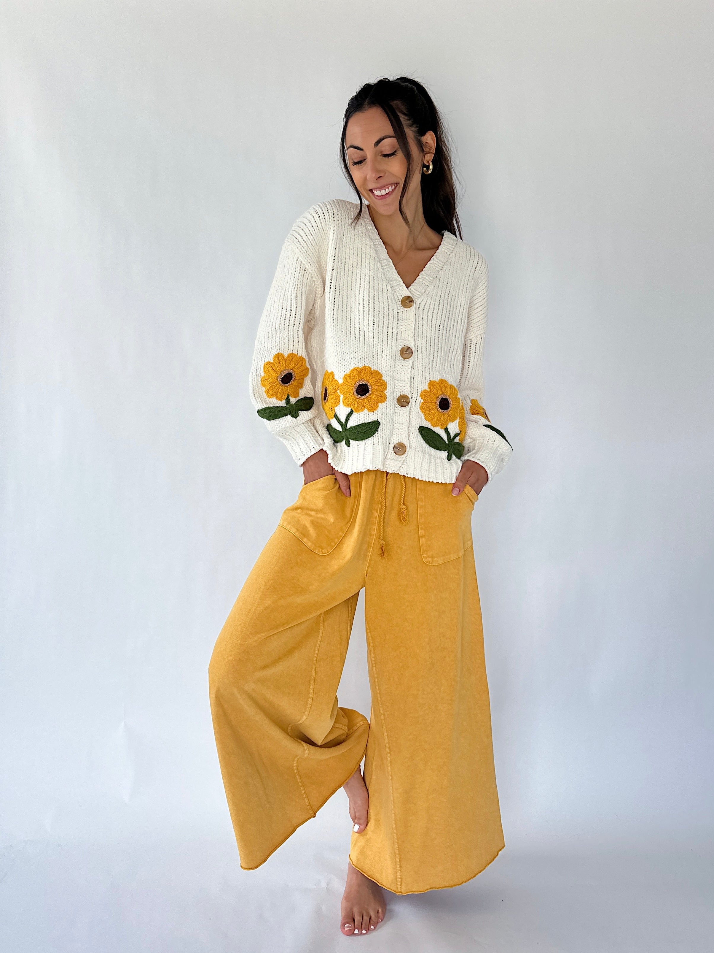 A woman modeling a cream ribbed cardigan with yellow sunflower embroidery on the front and sleeves, paired with mustard yellow wide-leg pants, against a plain white studio background.