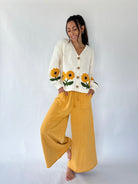A woman modeling a cream ribbed cardigan with yellow sunflower embroidery on the front and sleeves, paired with mustard yellow wide-leg pants, against a plain white studio background.
