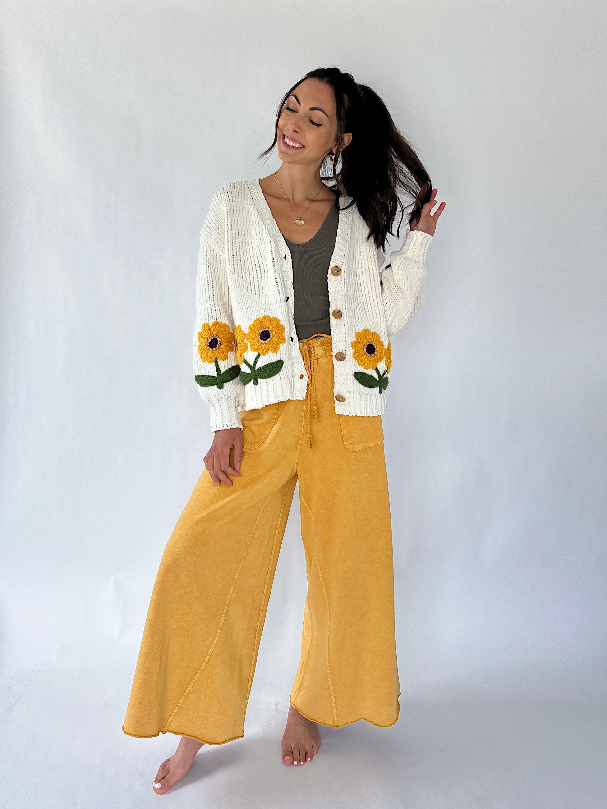 A woman modeling a white knit cardigan with yellow sunflower embroidery, paired with mustard yellow wide-leg pants and a dark gray top, against a plain white studio background.